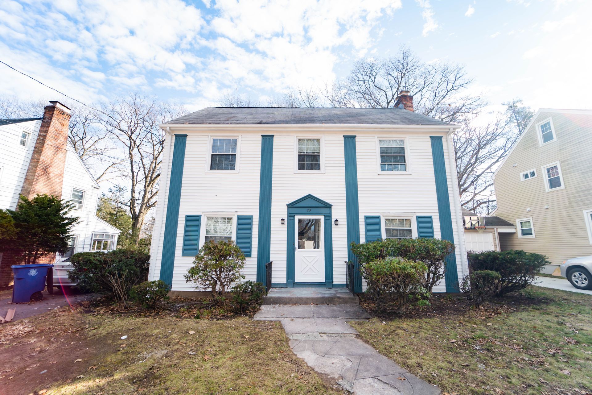 A two-story white house with dark teal trim, a centered front door, and manicured bushes in the front yard.