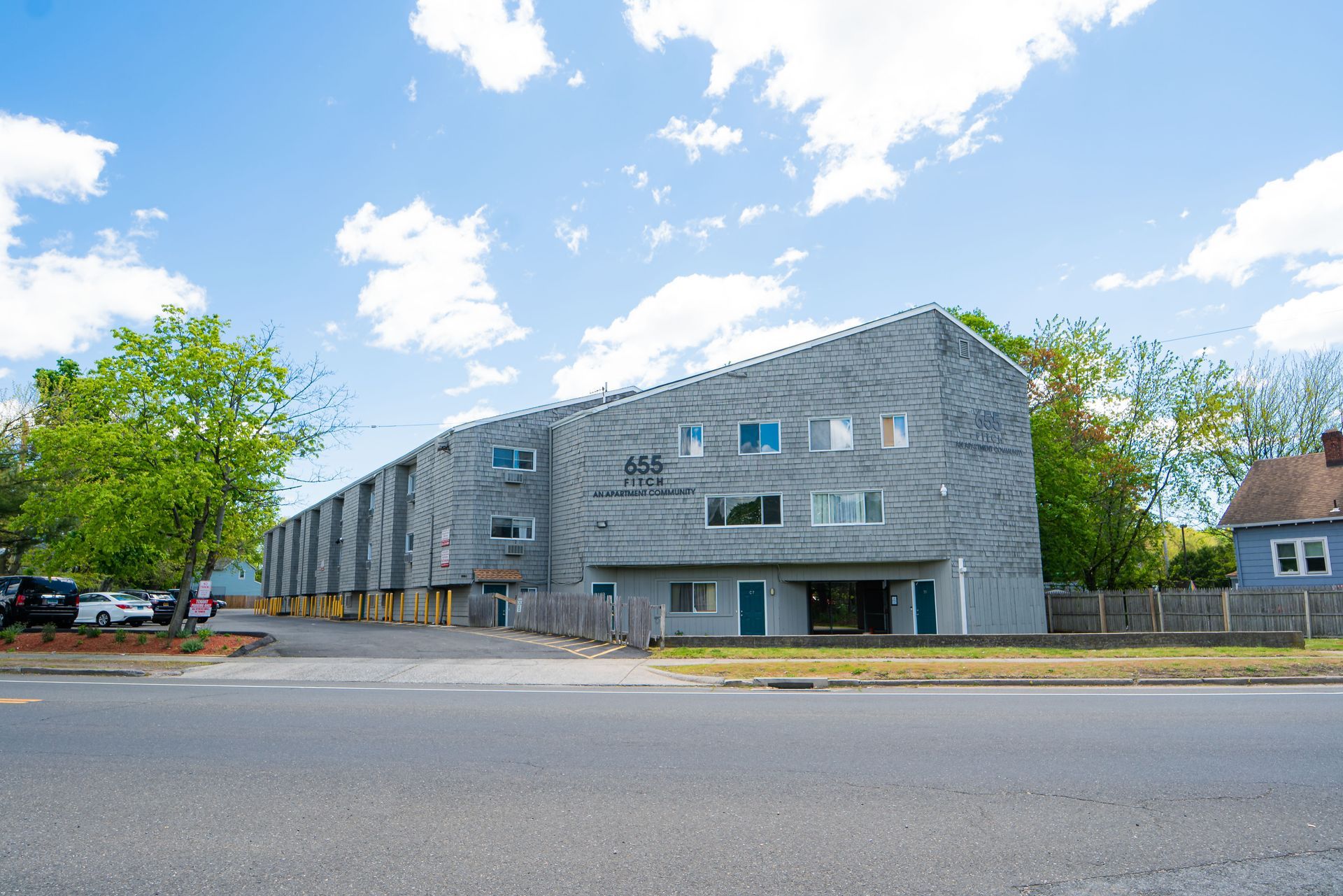 A three-story gray apartment building with a slanted roof, seen from across a street under a partly cloudy blue sky.