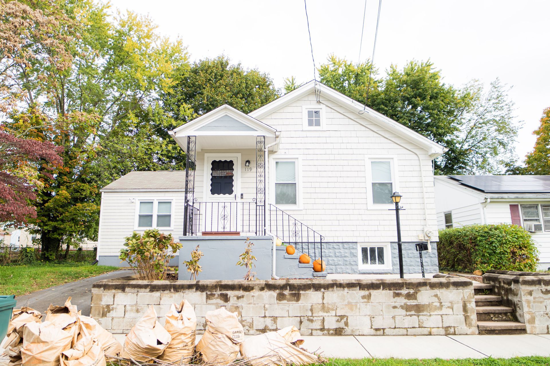 A white, one-story house with a small front porch, stone base, and several brown yard waste bags on the front lawn.