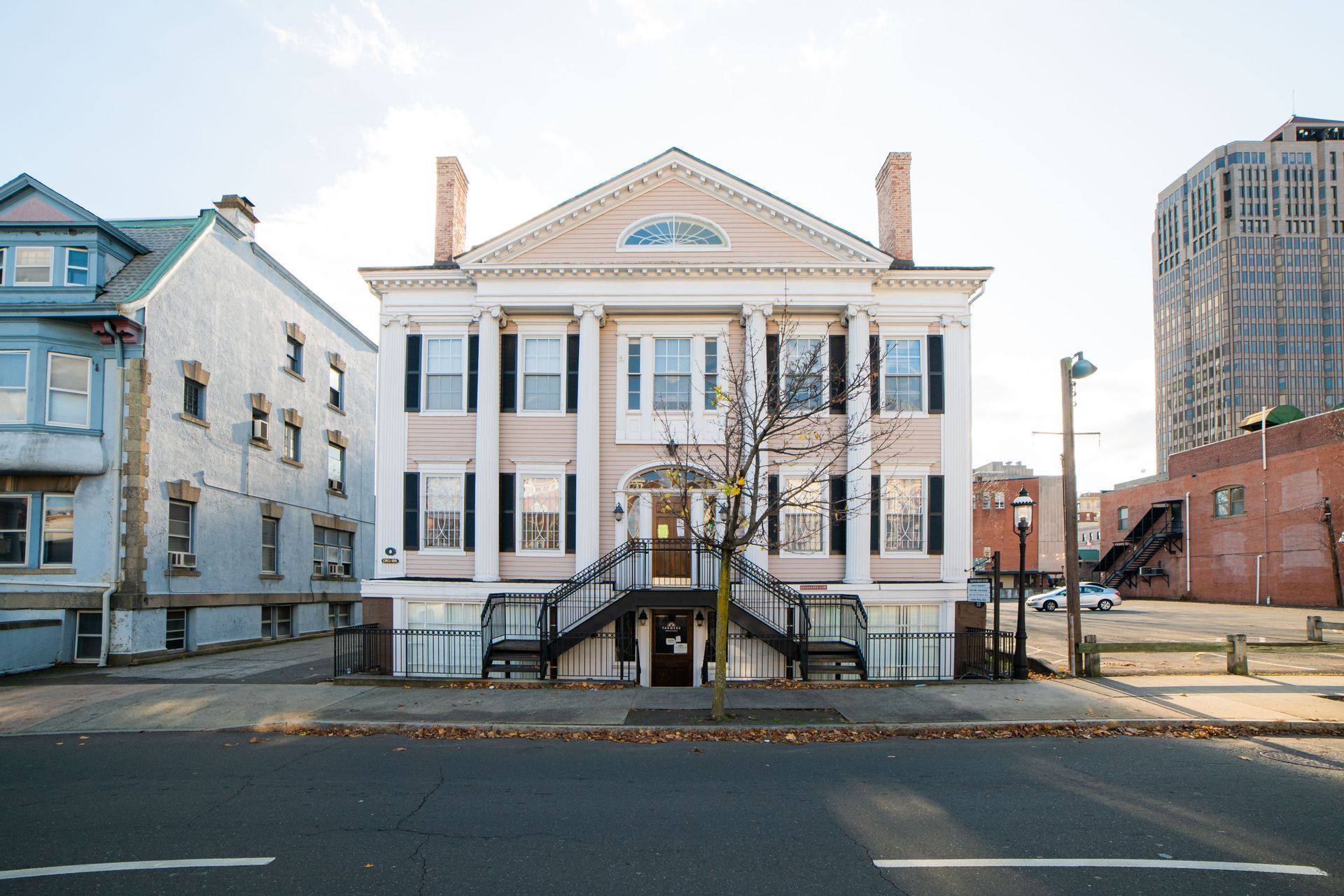 A pale pink, three-story building with white columns and a central staircase, set between other buildings on a city street.