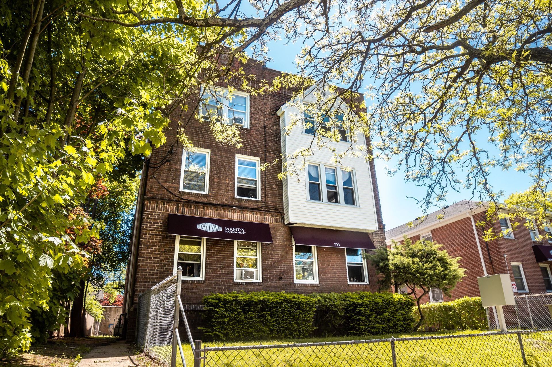 A three-story, brown brick apartment building with white siding and dark awnings, surrounded by trees on a sunny day.