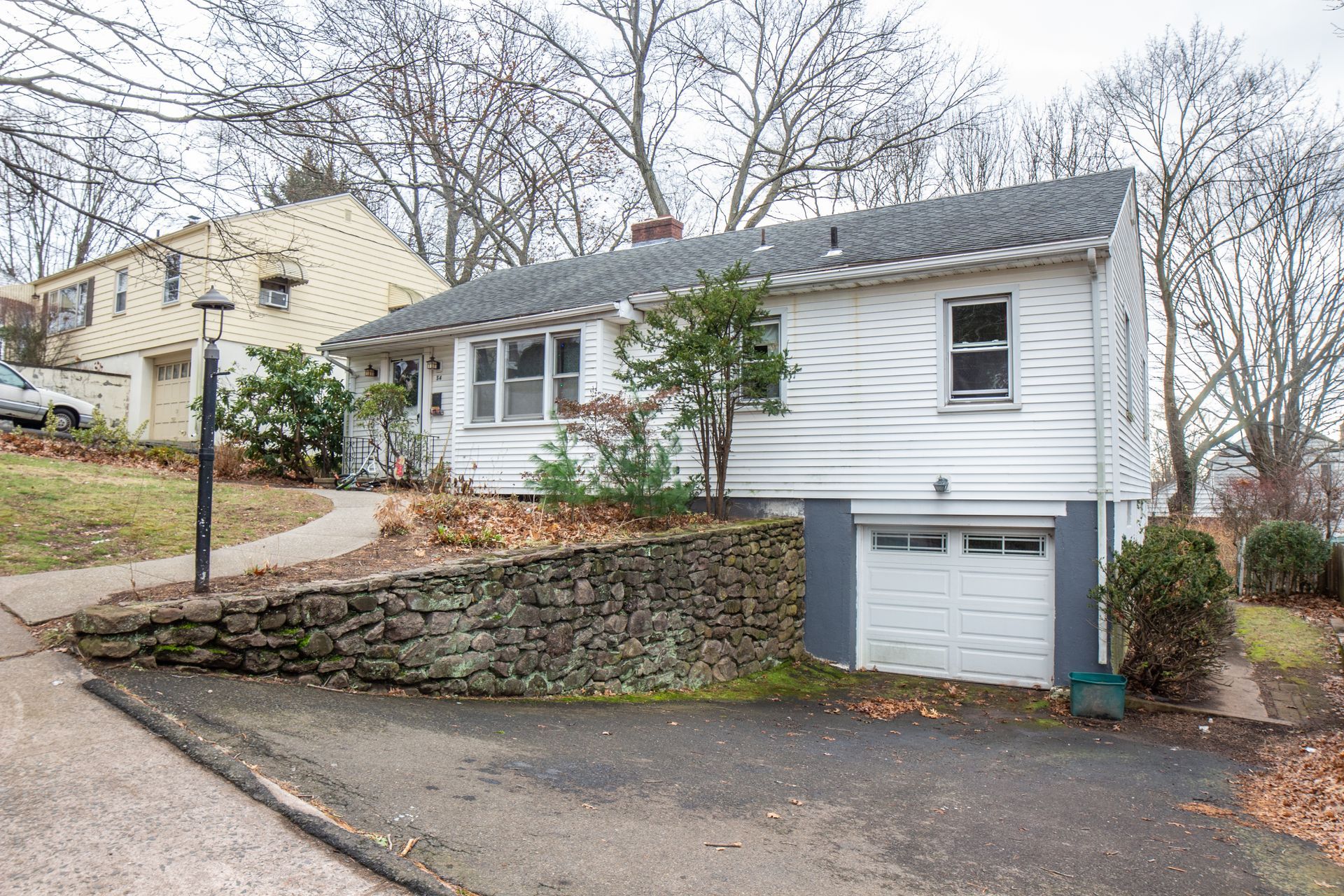 A white, split-level house with a stone retaining wall, a paved driveway, and an integrated garage on a sloped lot.