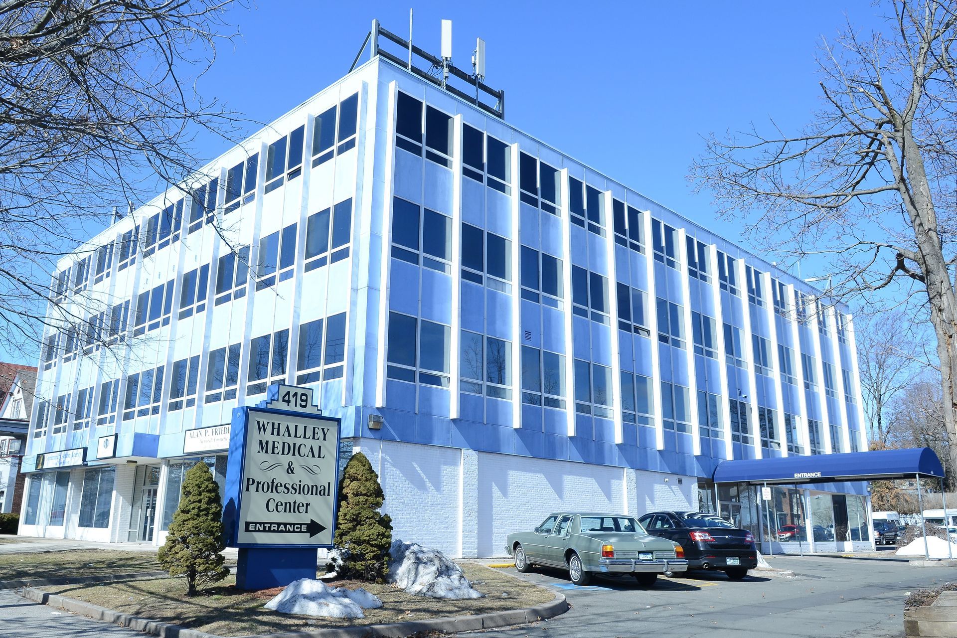 A multi-story blue office building with a directional sign and parked cars in front under a clear sky.
