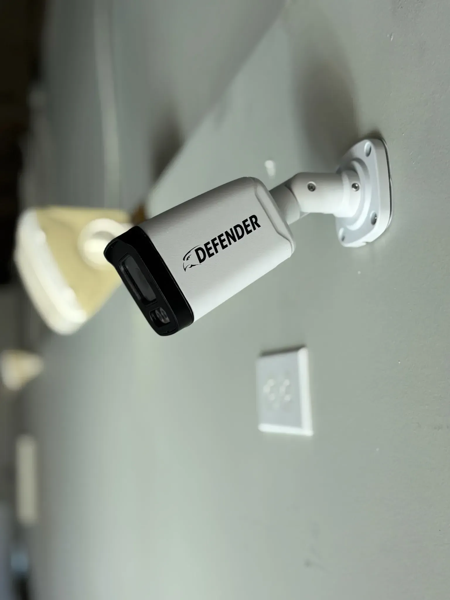 A white Defender surveillance camera mounted on a plain, light-colored wall.