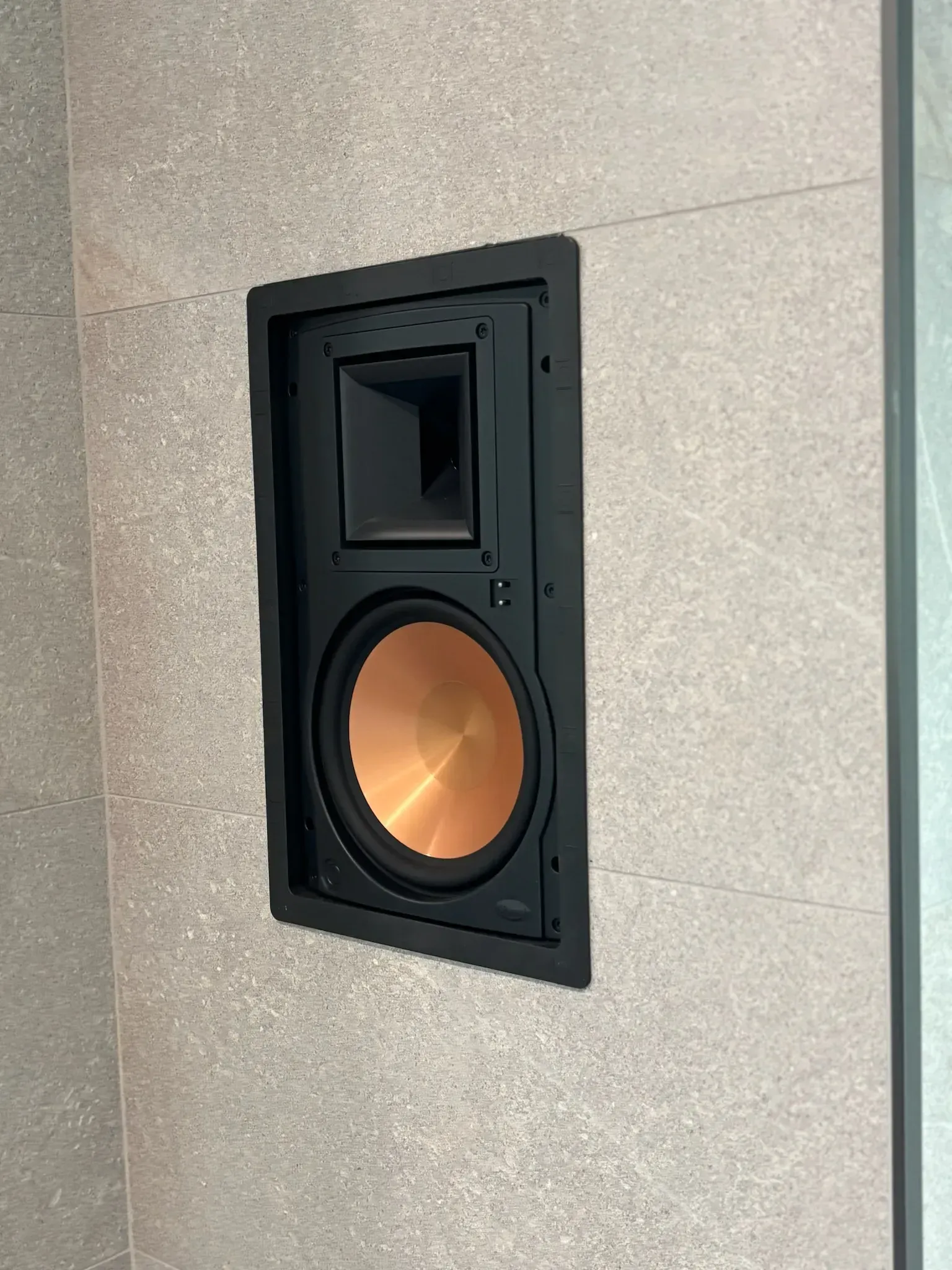 A rectangular Klipsch in-wall speaker with a black frame and a distinct copper-colored woofer mounted in a stone wall.