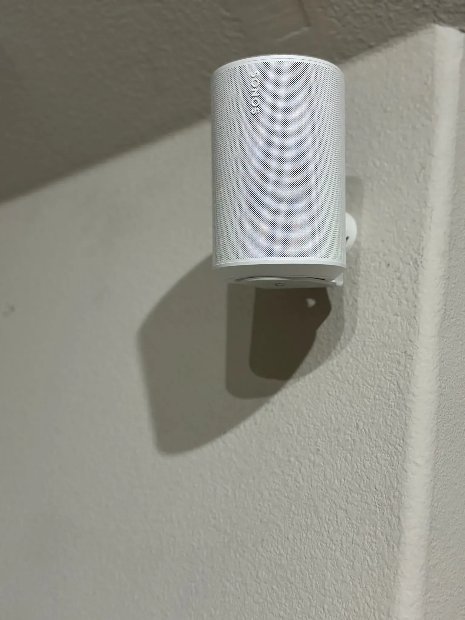 A white, cylindrical wall-mounted light fixture with a wavy, patterned mesh cover, casting a shadow on a textured wall.
