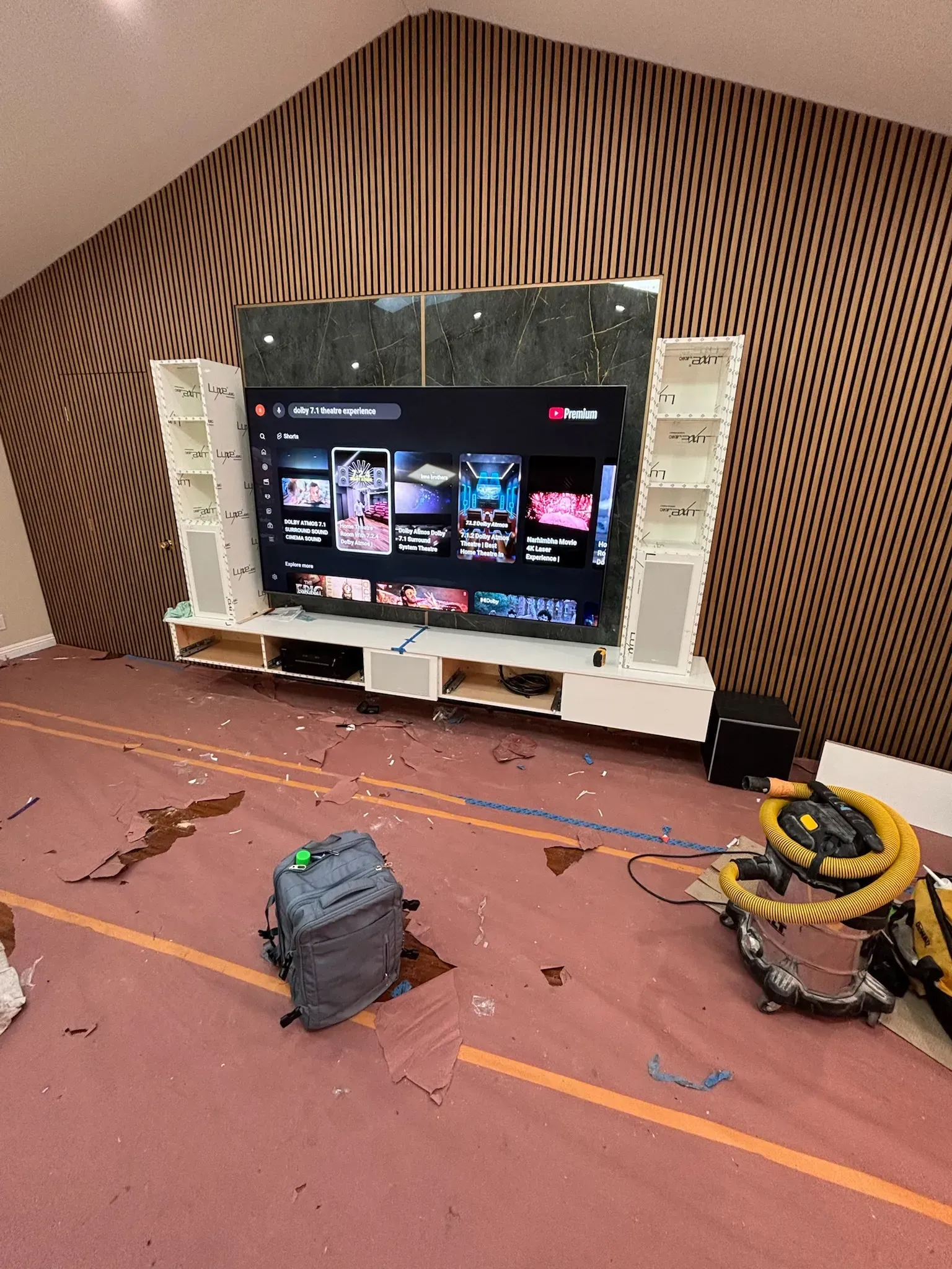 A wall-mounted TV unit with shelving sits against a patterned wallpaper, with debris and a shop vacuum on the floor.
