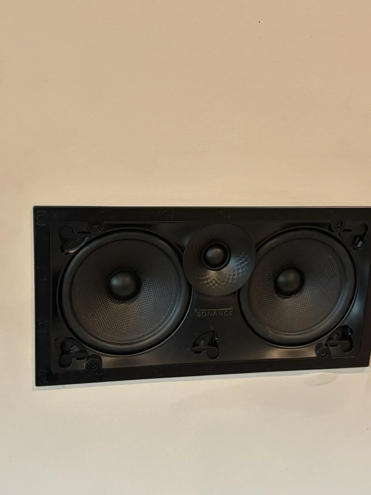 A rectangular wall-mounted speaker with two black woofers flanking a central tweeter, set against a plain beige wall.