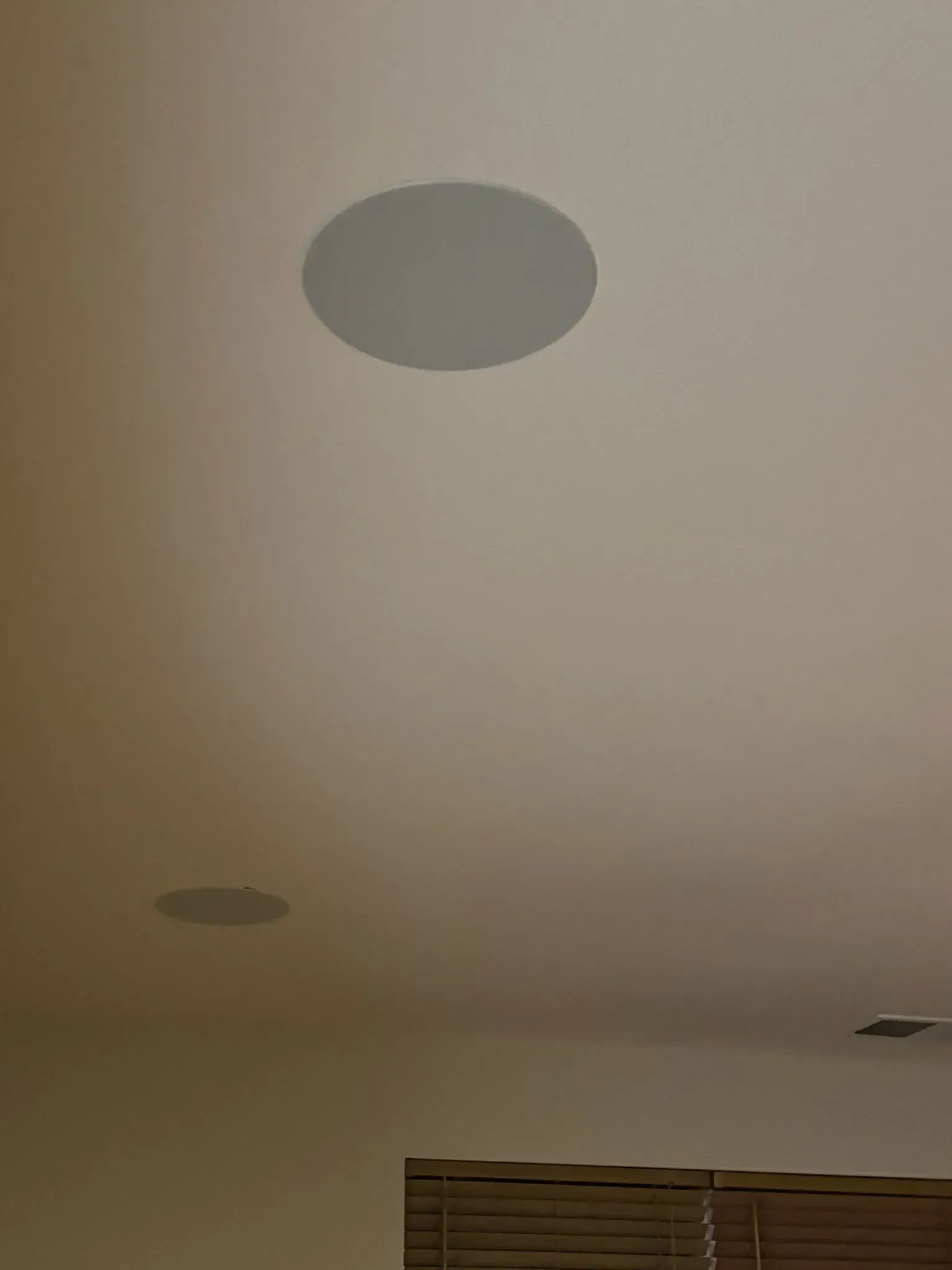 Two round, white speakers are mounted in a plain, light-colored ceiling above window blinds.