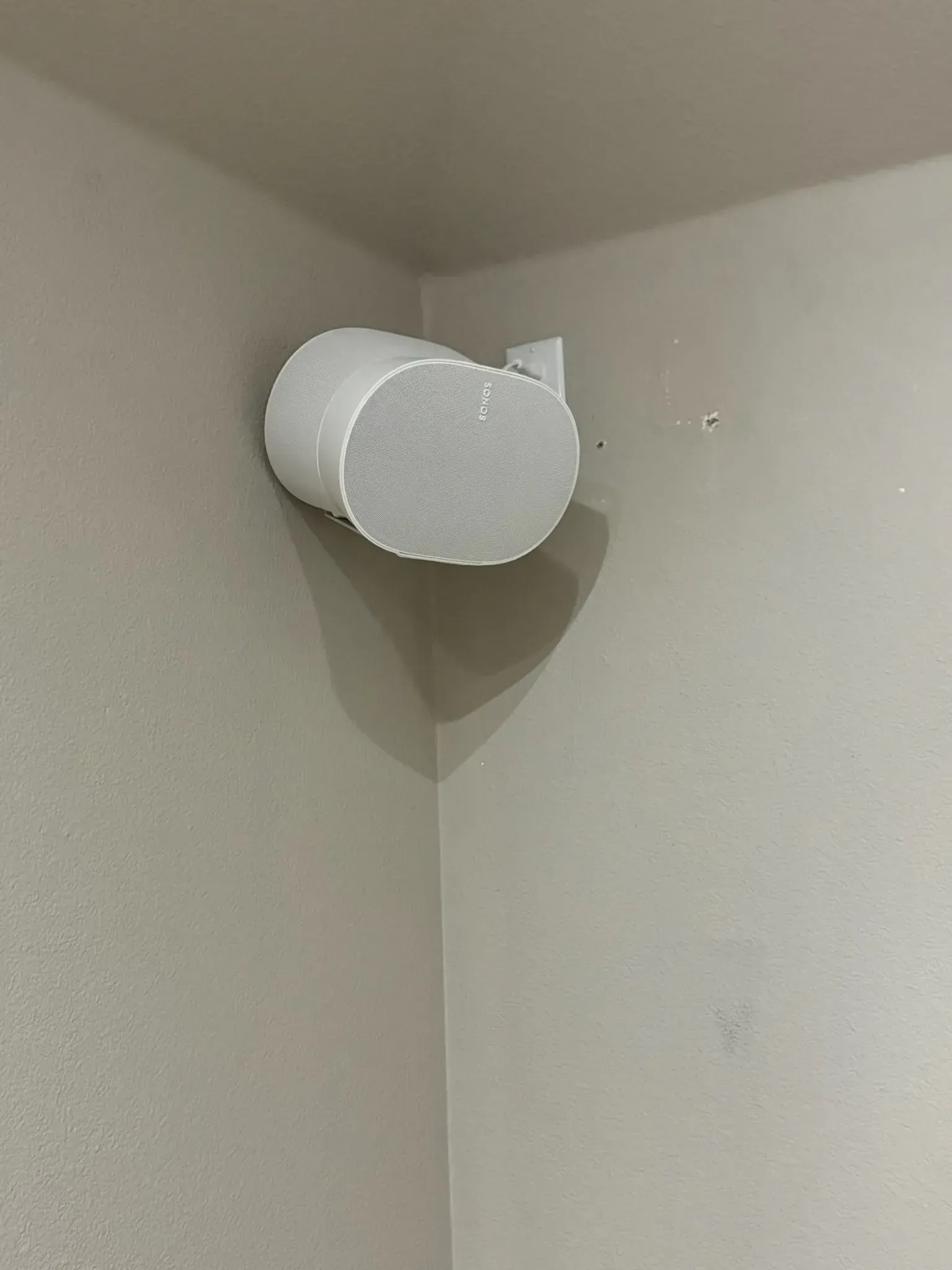 A white, oval-shaped smart speaker mounted in the corner of a beige wall.