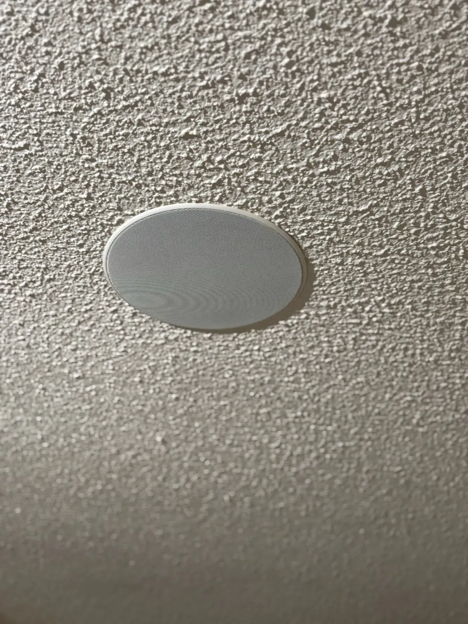 A round, white recessed ceiling speaker with a decorative mesh grille, mounted in a textured, off-white popcorn ceiling.