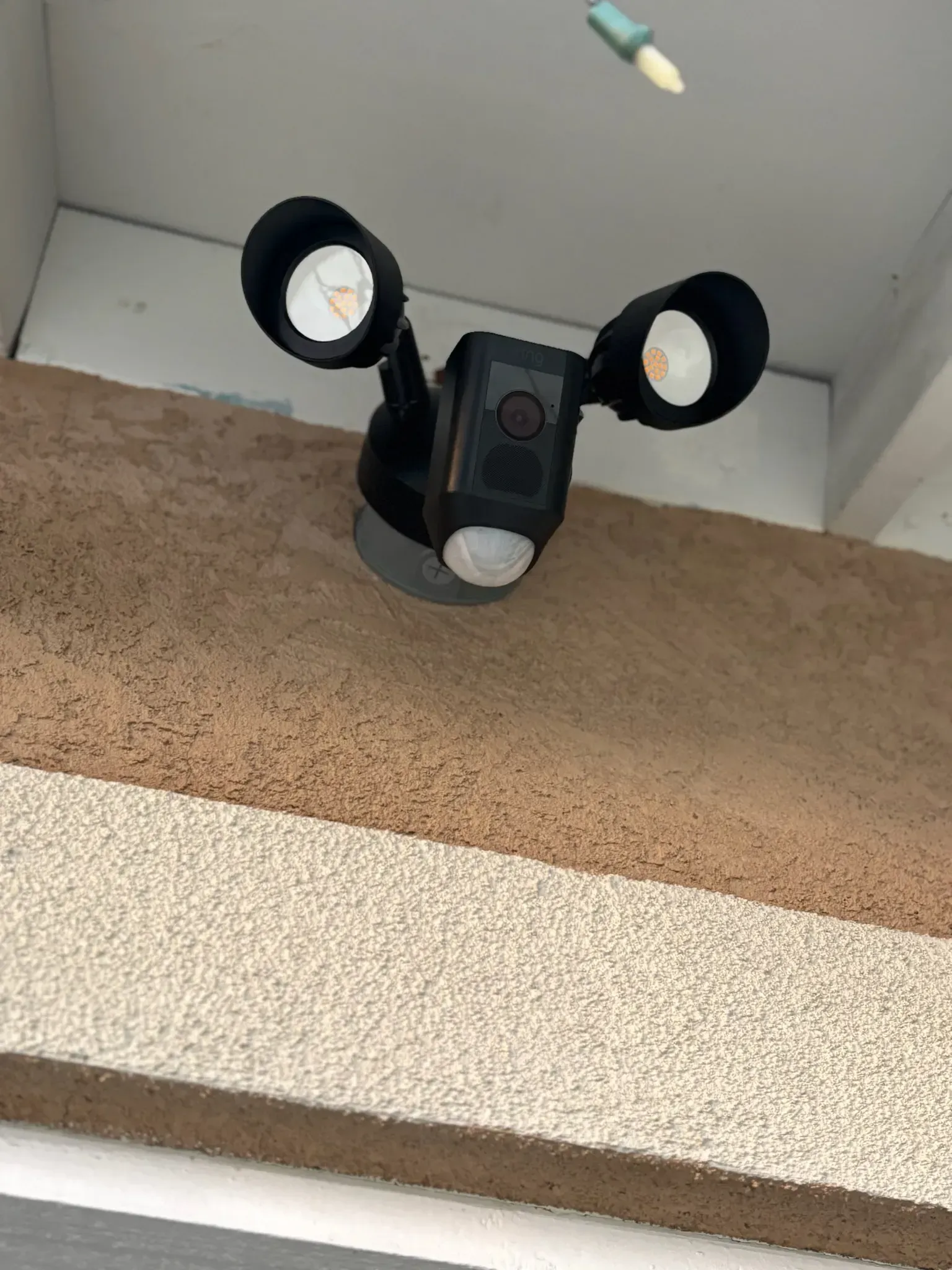 A black outdoor security floodlight with two adjustable LED lamps and a built-in camera mounted on a textured wall.