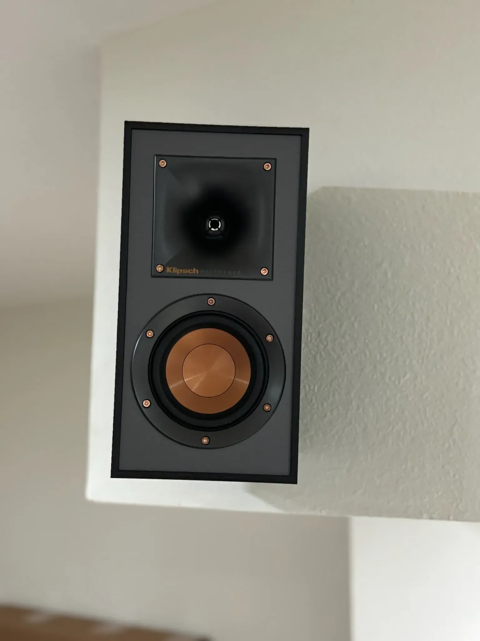 A black Klipsch bookshelf speaker with a signature copper-colored woofer and horn tweeter, mounted on a white wall corner.