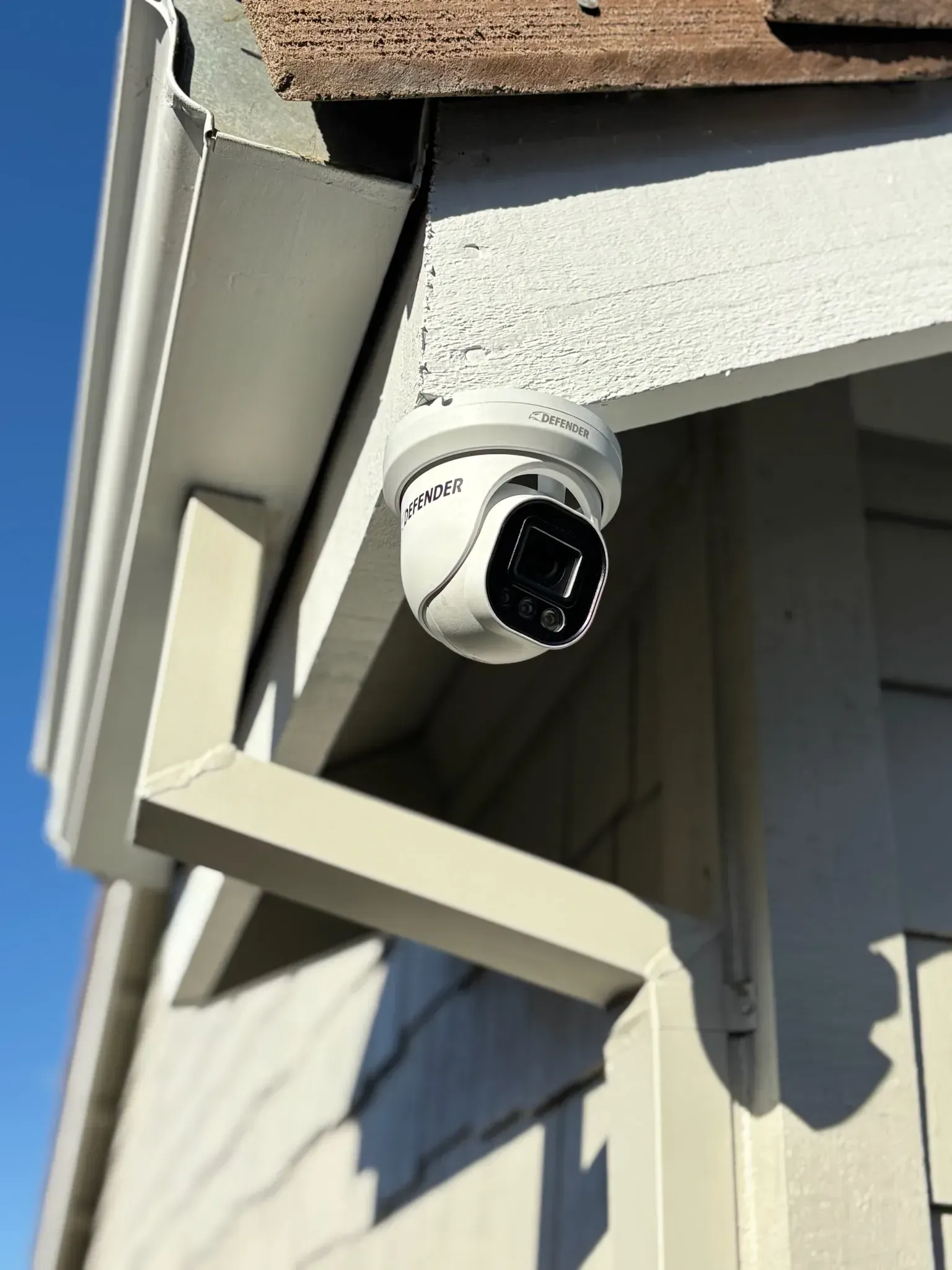 A white dome-style security camera mounted under the eaves of a light-colored house exterior.