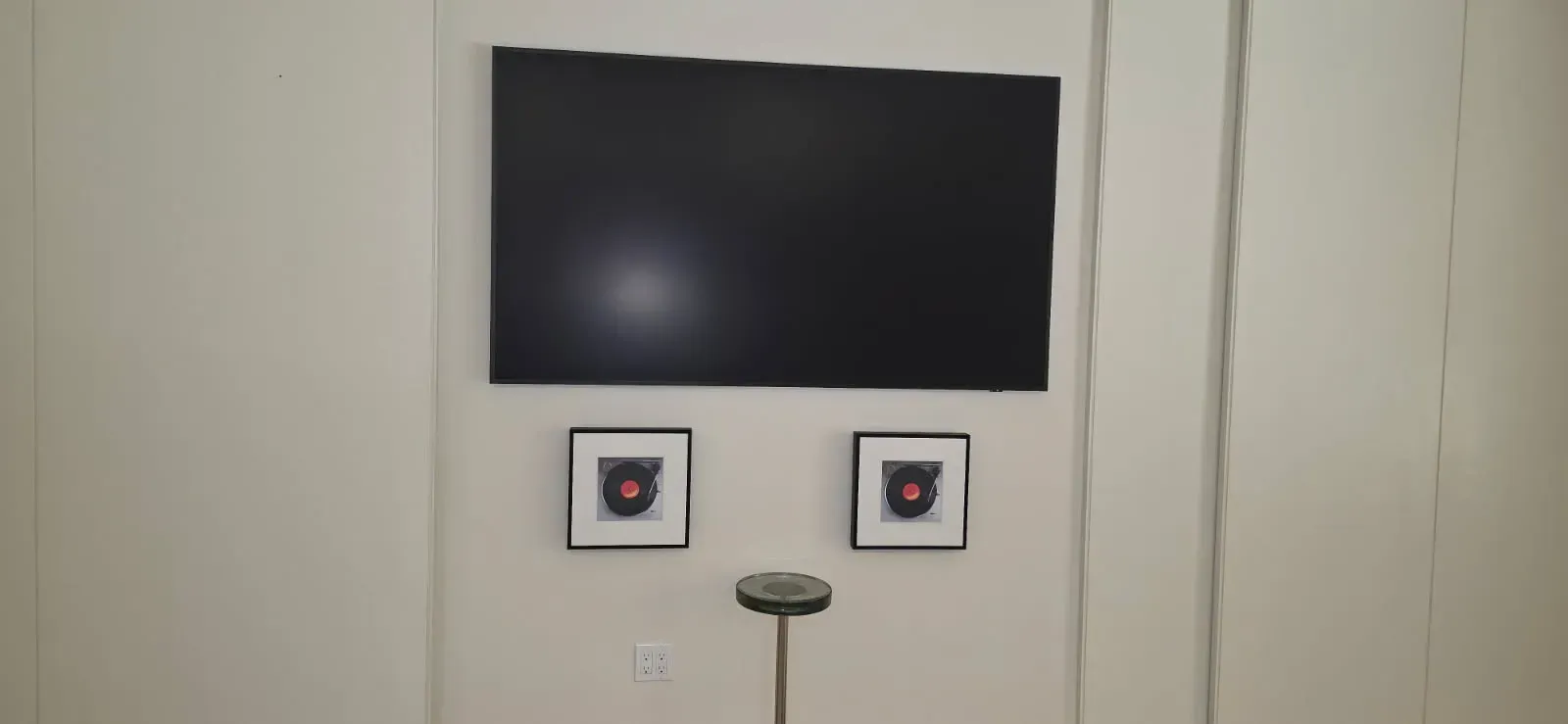 A wall-mounted flat-screen television above two framed square artworks and a small pedestal stand.