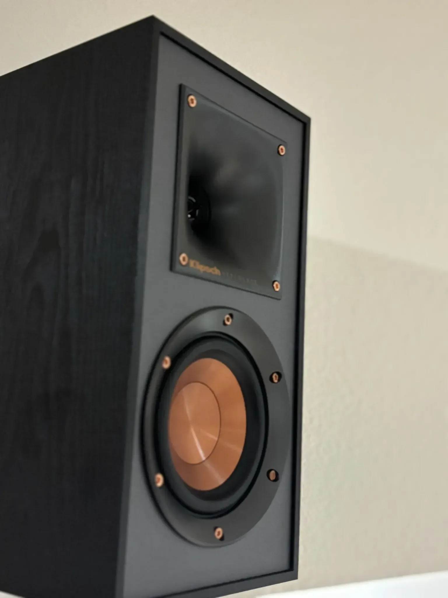 A black bookshelf speaker with a copper-colored woofer and a rectangular horn-loaded tweeter against a plain wall.