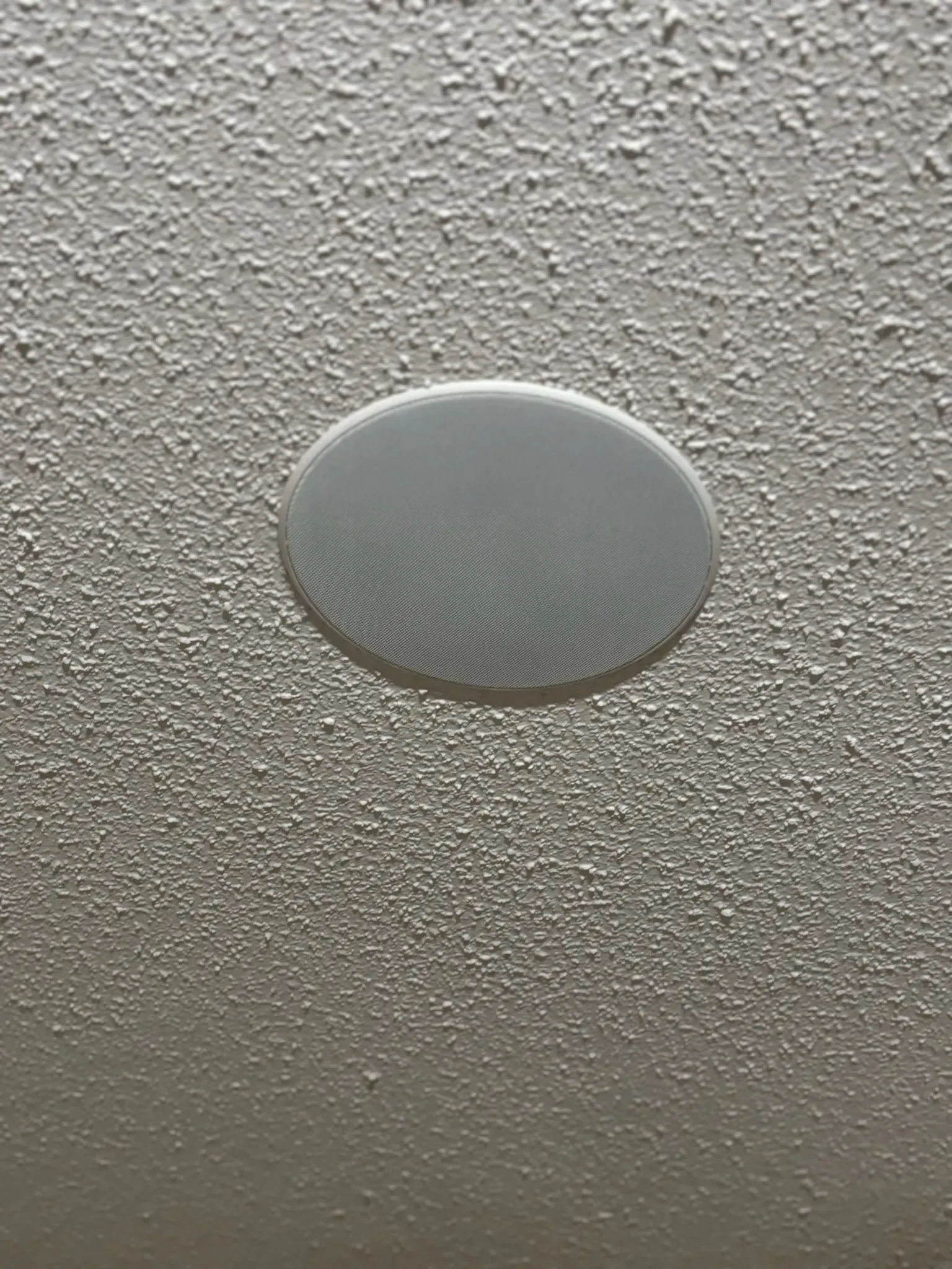 A round, white speaker mounted flush into a textured, white ceiling.