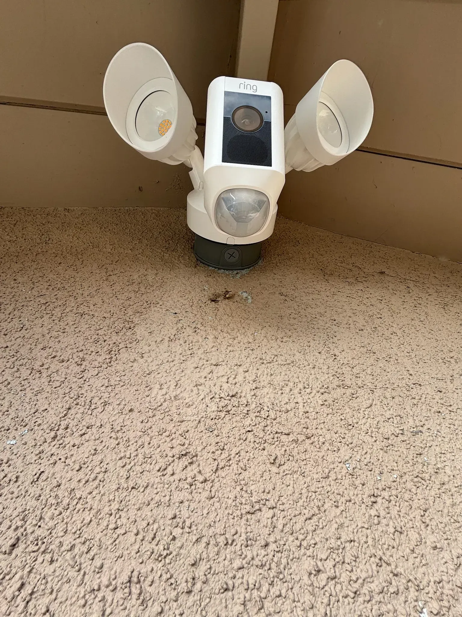 A white Ring floodlight security camera mounted on an interior wall above beige carpet.