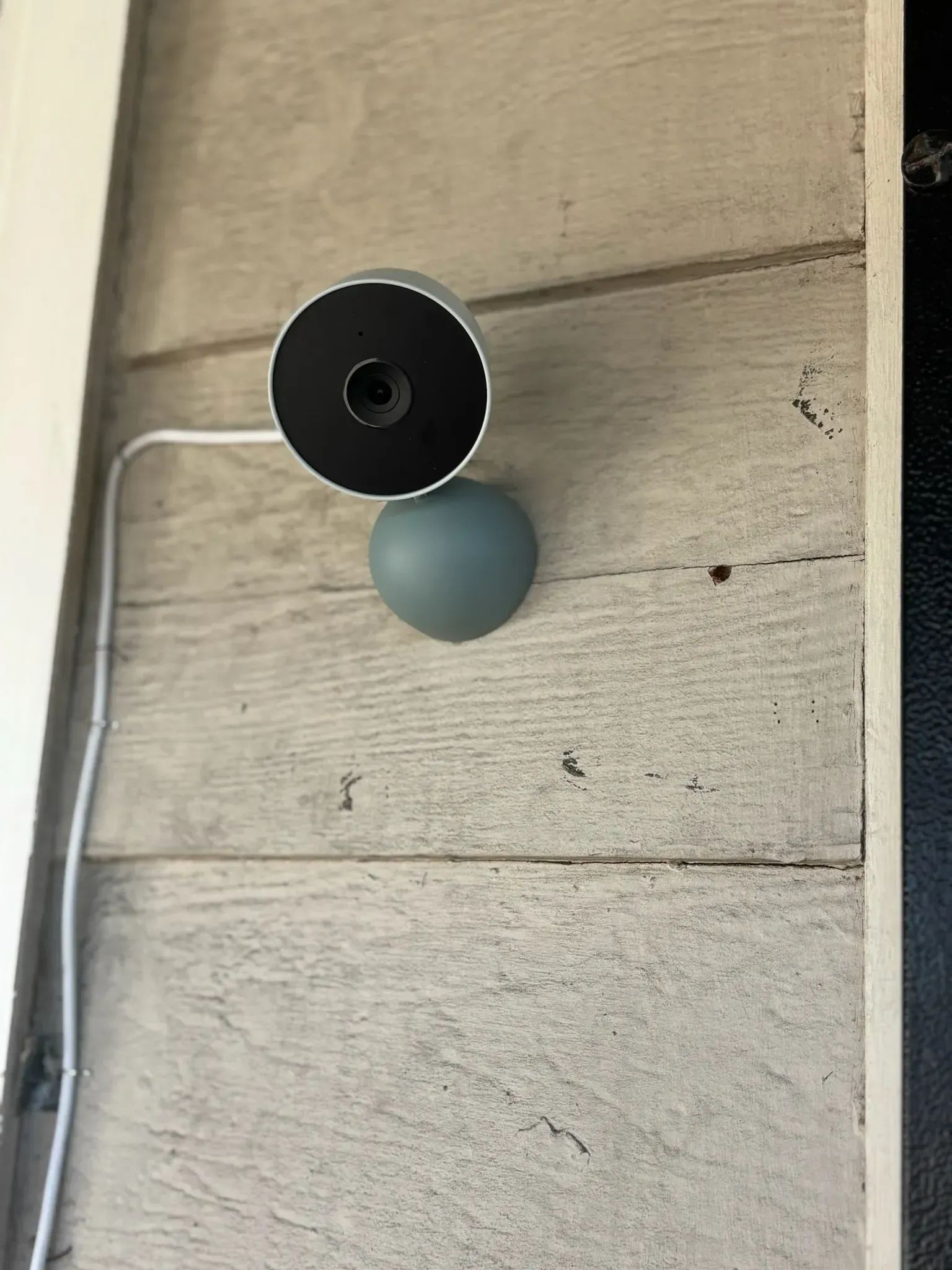 A gray, circular security camera mounted on a textured, light-colored exterior wall, with a white power cord extending out.