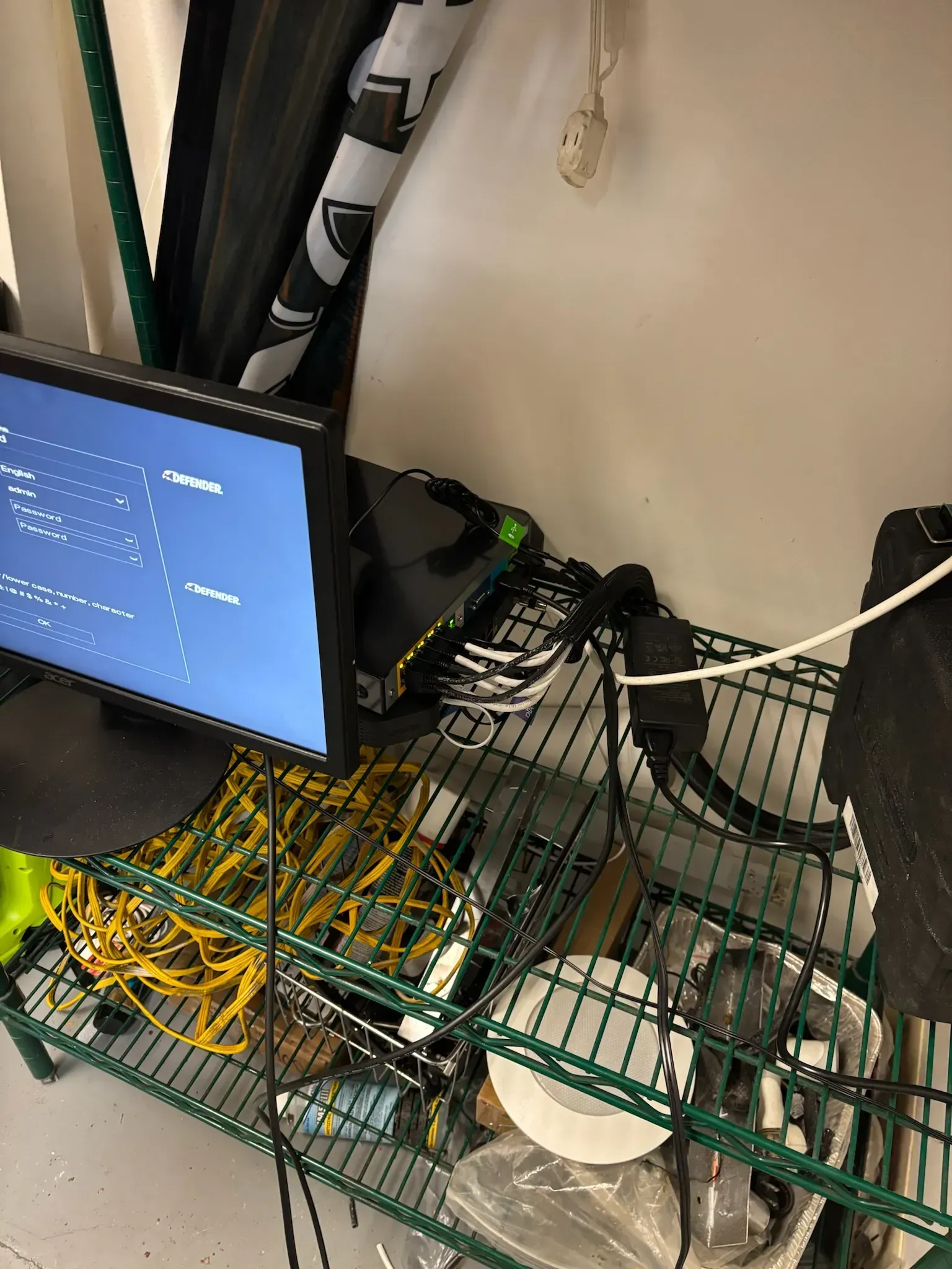 A green wire shelf holds a computer monitor displaying a blue screen, cables, and various electronic equipment.