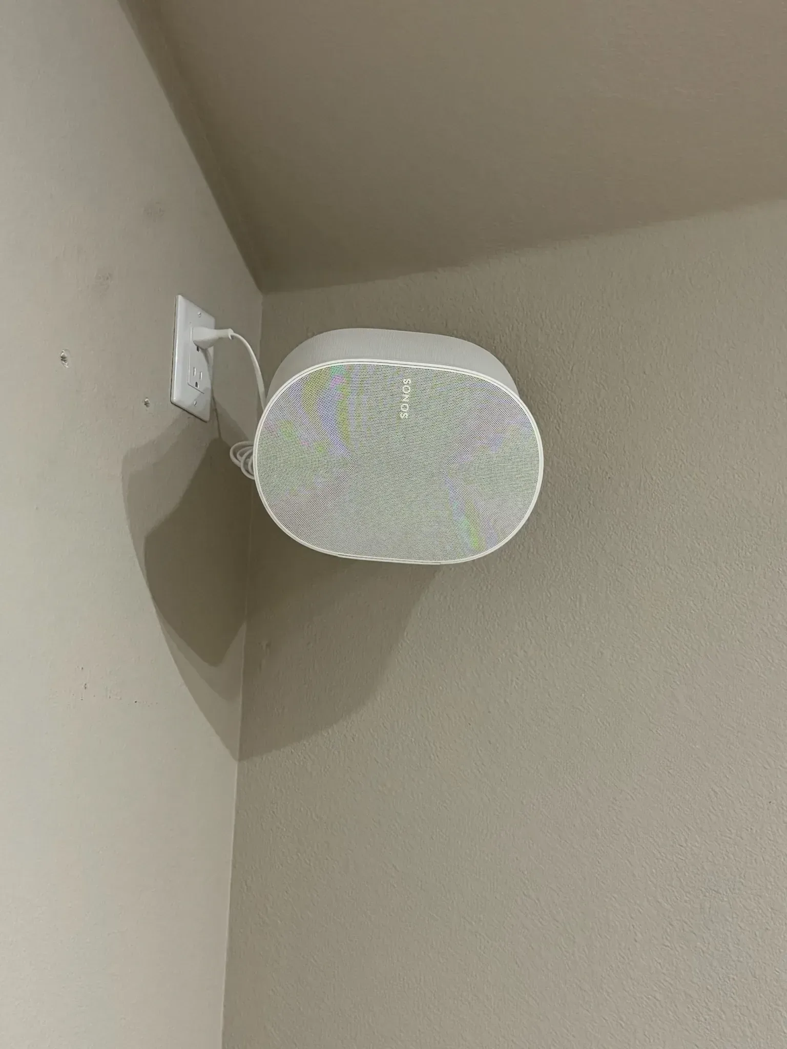 A white, oval-shaped smart speaker mounted on a corner wall, plugged into a power outlet.