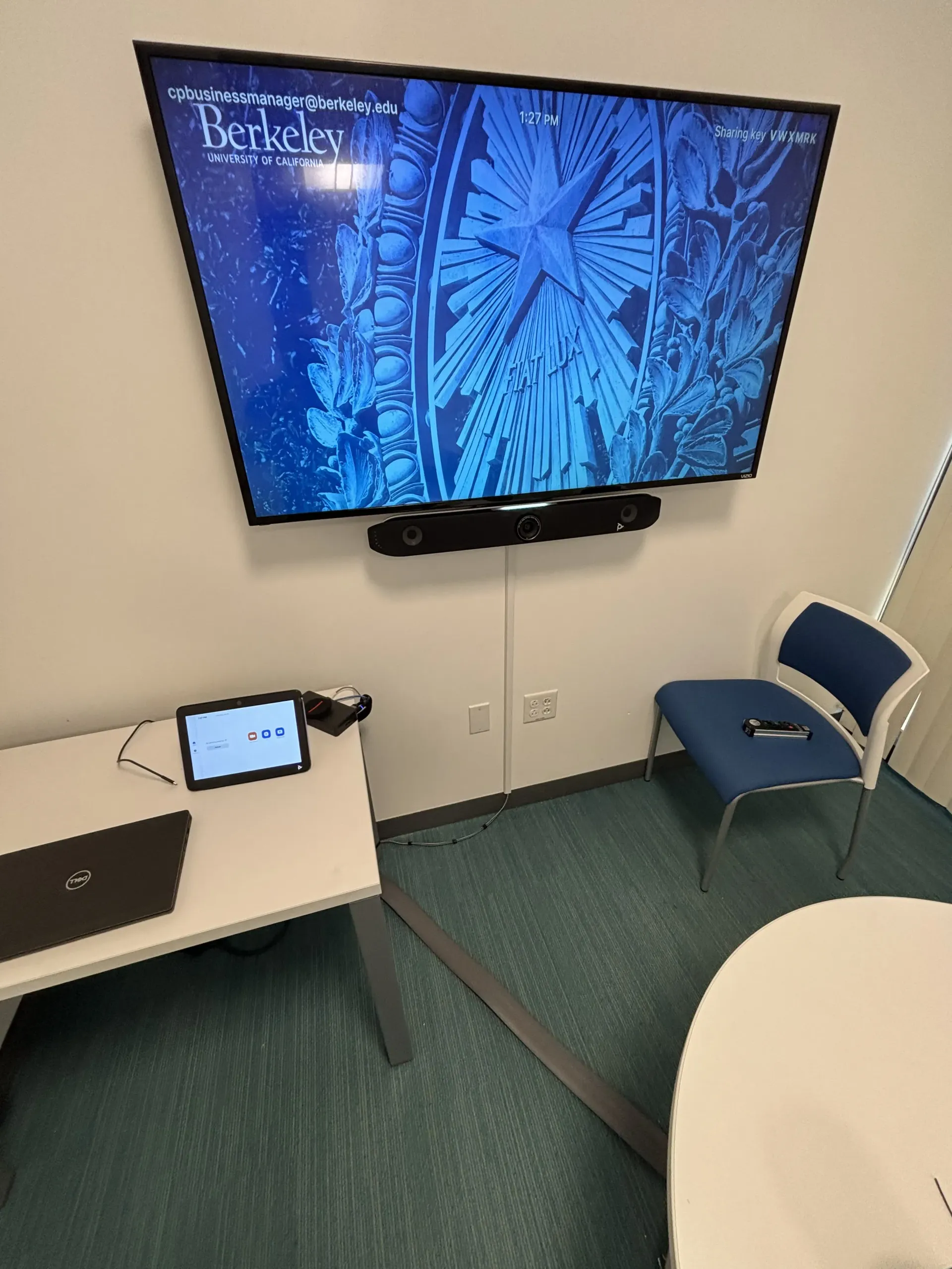A meeting room with a large wall-mounted monitor showing a blue University of California, Berkeley graphic, a desk, and chair.