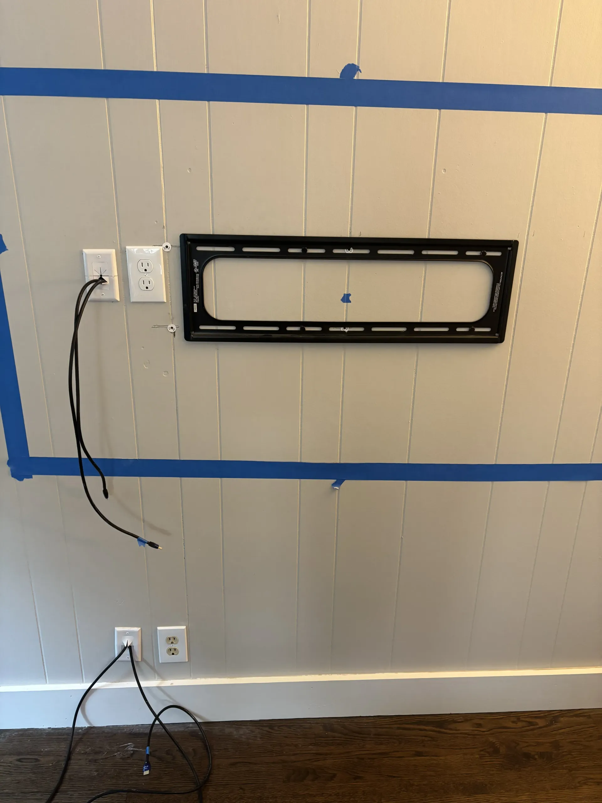 A wall-mounted TV bracket on a light-colored wood panel wall with exposed power cables and blue painter's tape markings.