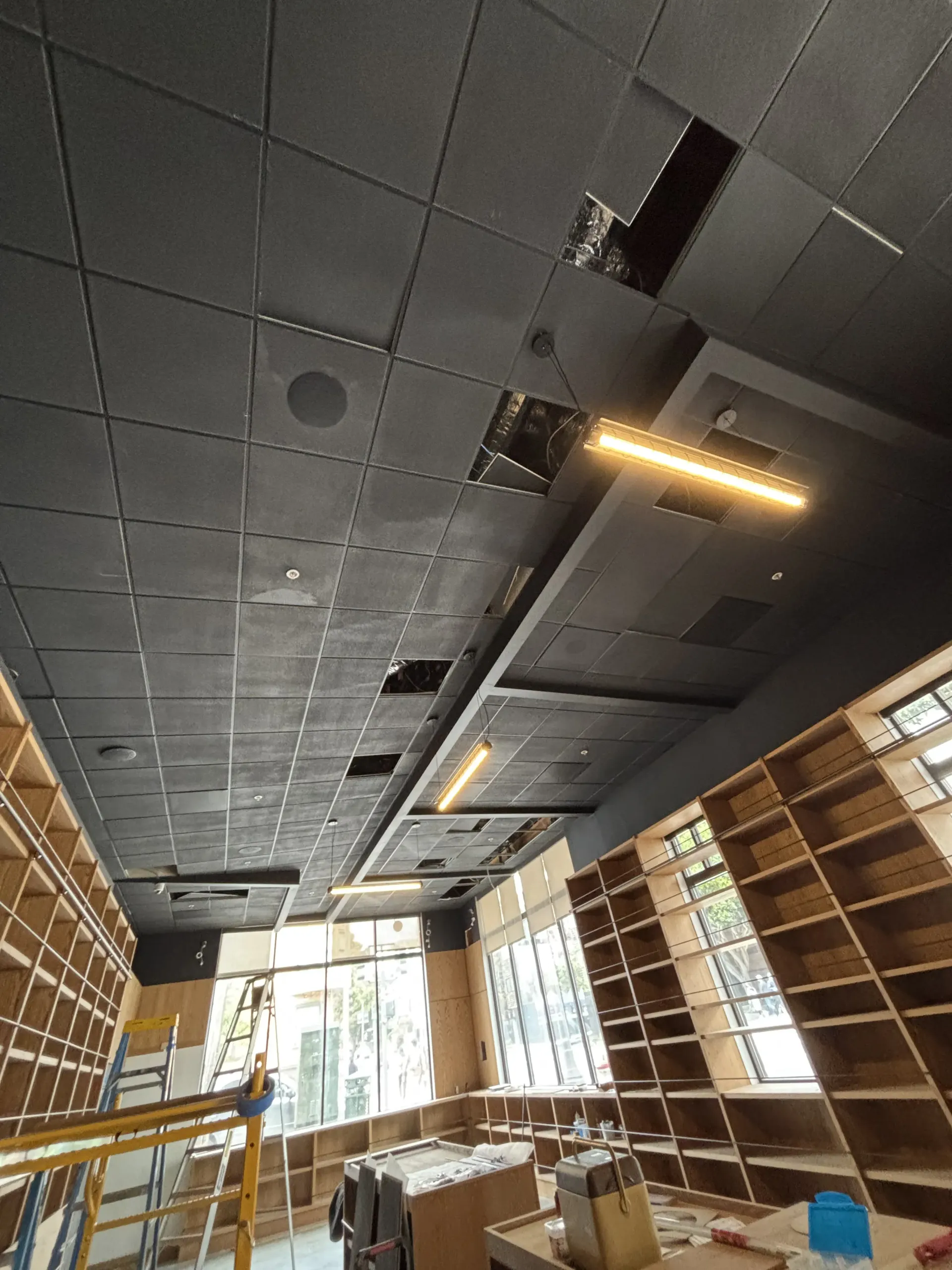 An interior view of a room under construction, featuring wooden shelving units and a black grid ceiling with missing panels.