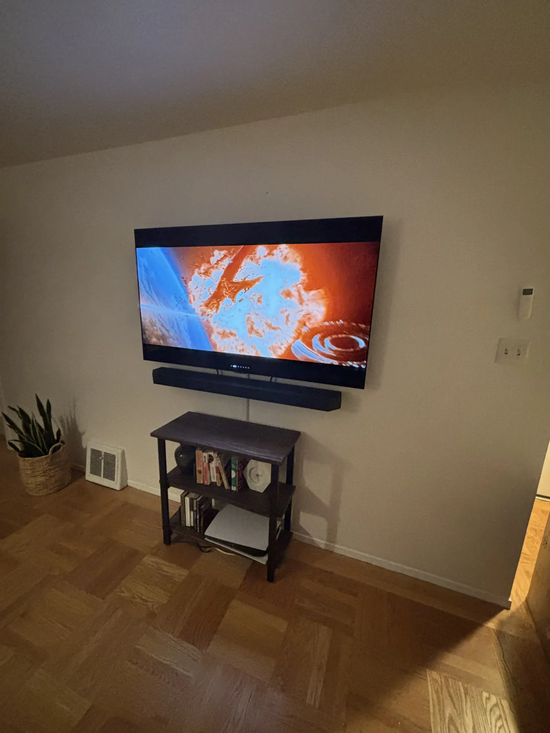 A wall-mounted TV playing a bright, orange-toned video above a soundbar and a three-tiered shelf on a wooden floor.