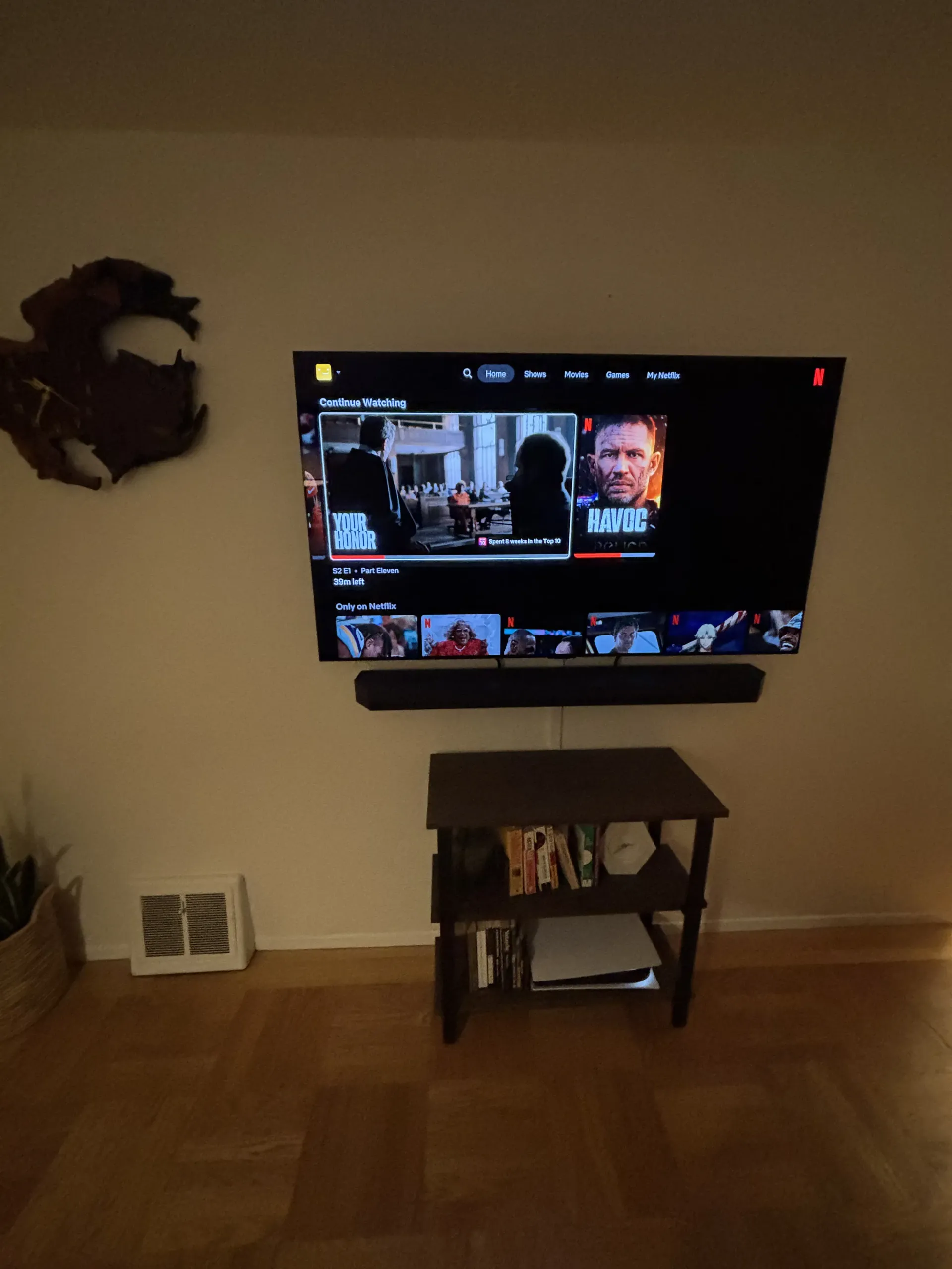 A wall-mounted TV showing a movie interface above a small wooden shelf with a gaming console, with wall art to the left.