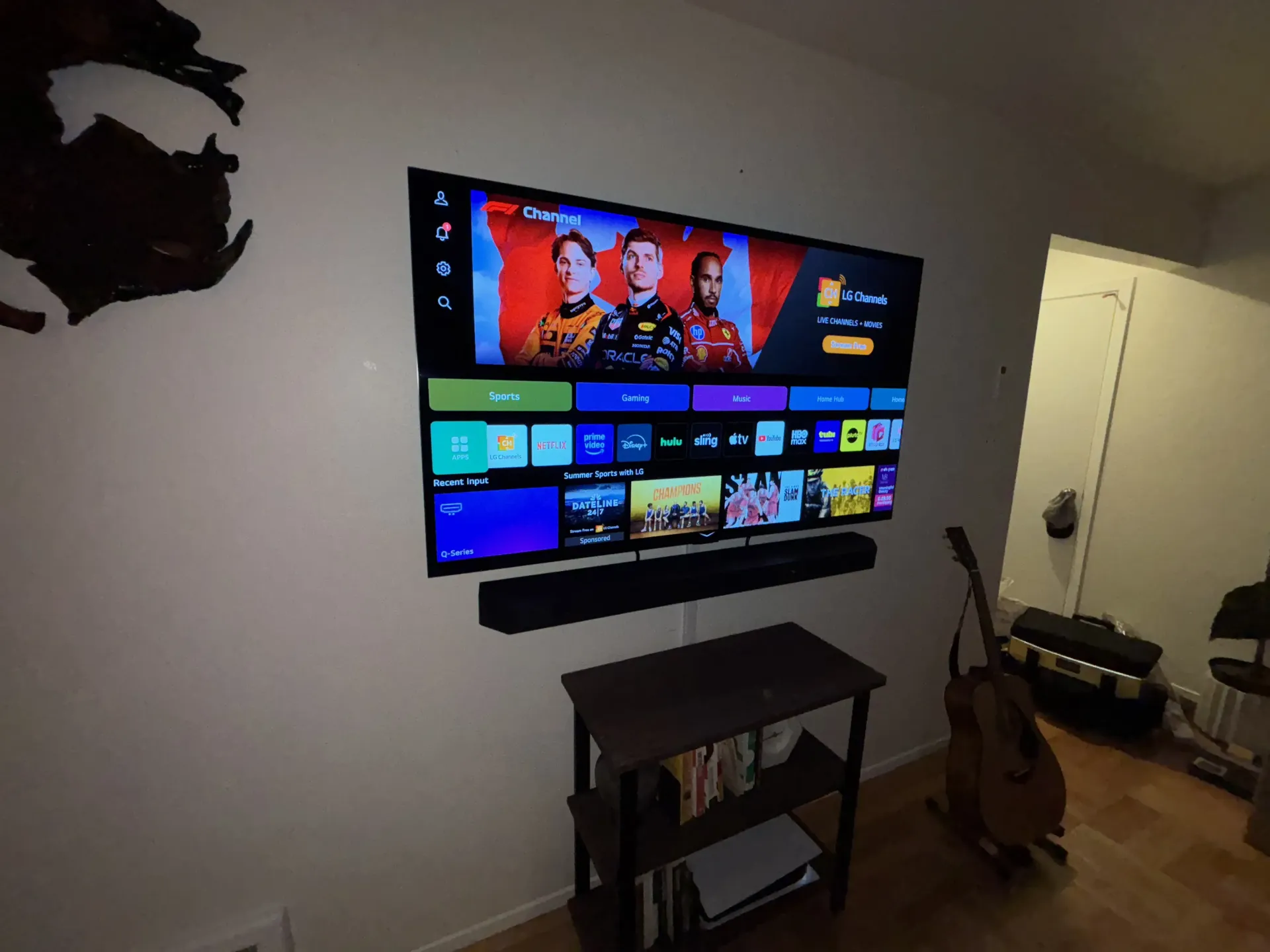 A large wall-mounted flat-screen TV displaying a streaming menu sits above a soundbar and a small table with a guitar.