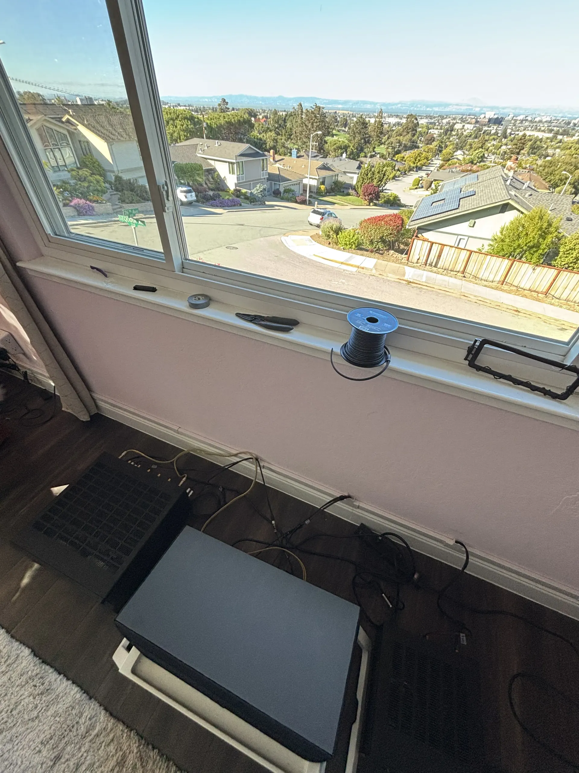 A window ledge holds a spool of wire, pliers, and small tools, overlooking a suburban street with houses and trees.