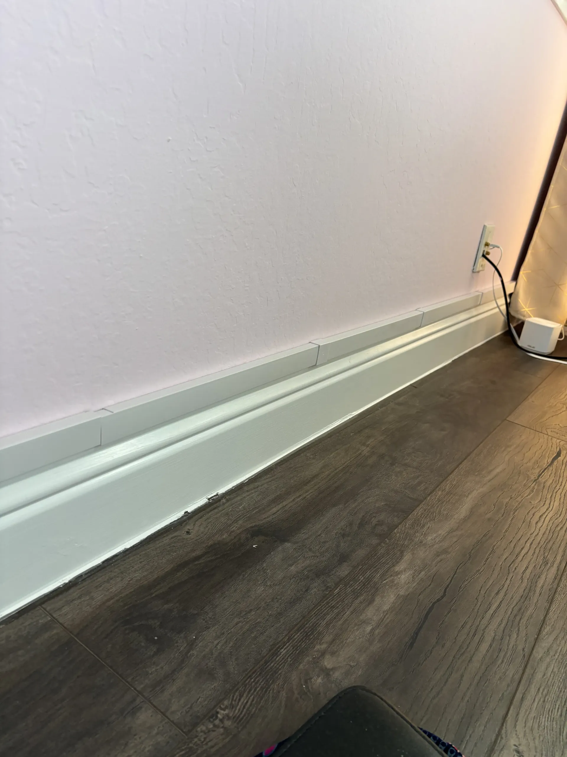 A view of a painted baseboard meeting dark wood flooring, with a wall outlet visible in the background.