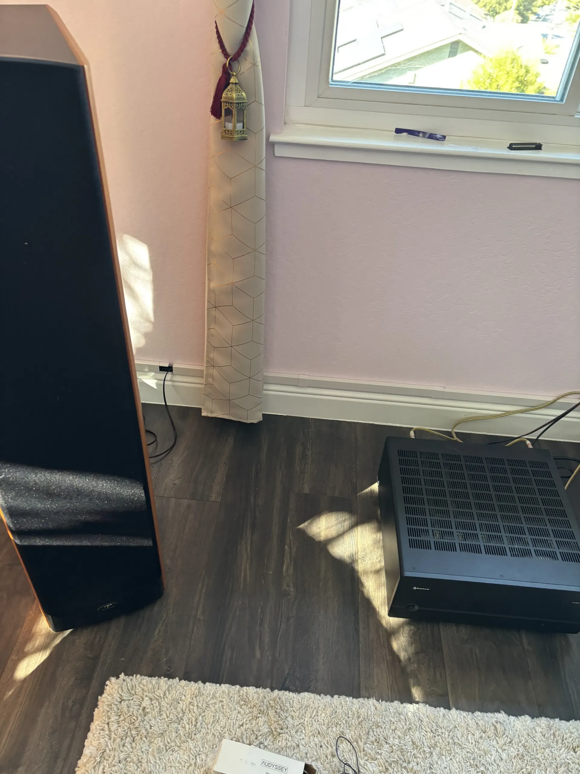 A tall black floor speaker stands beside a black patterned metal grate on a wood-look floor near a window.