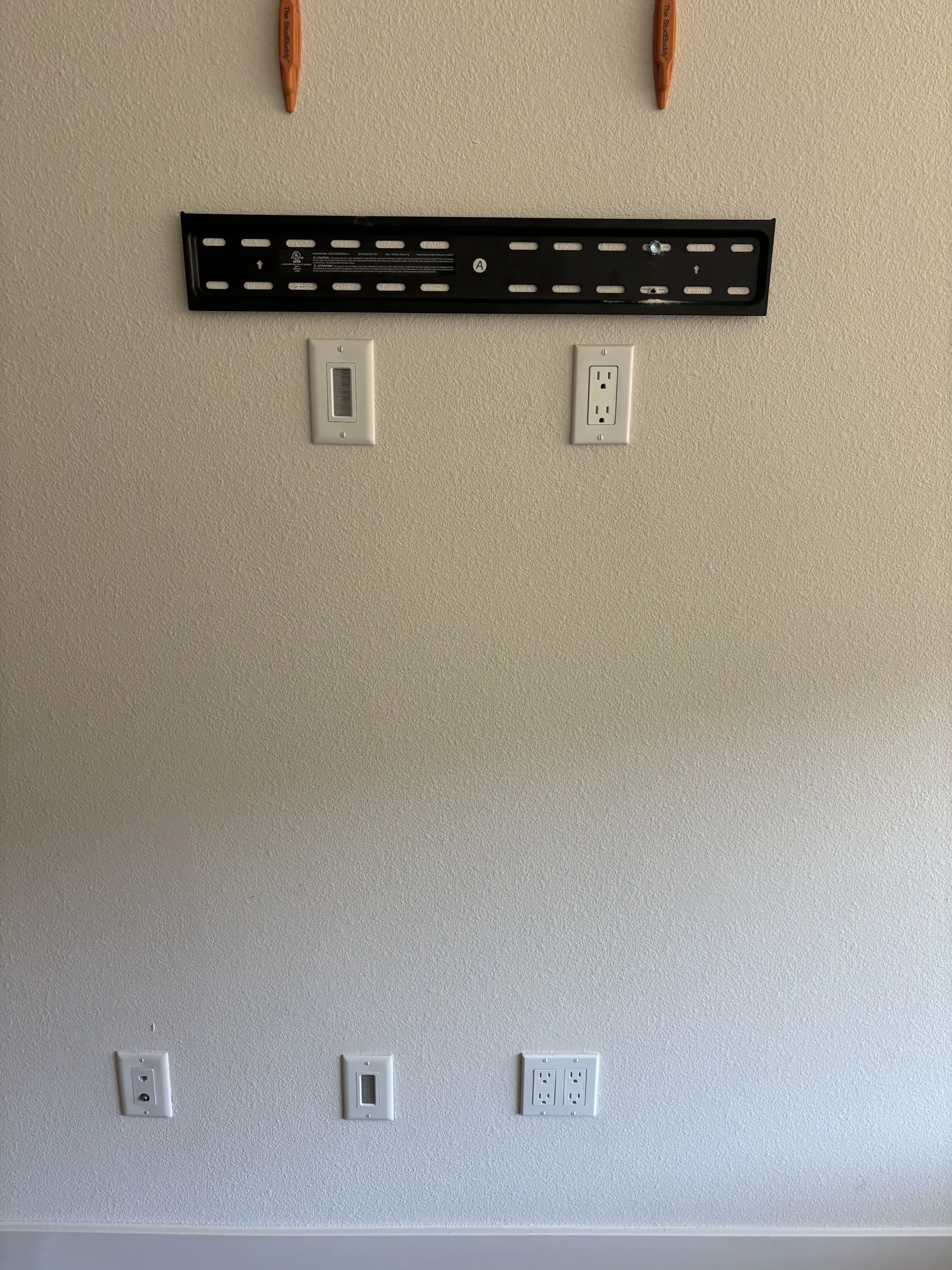 A wall with a mounted TV bracket, an upper power outlet and cable pass-through, and lower outlets and cable ports.