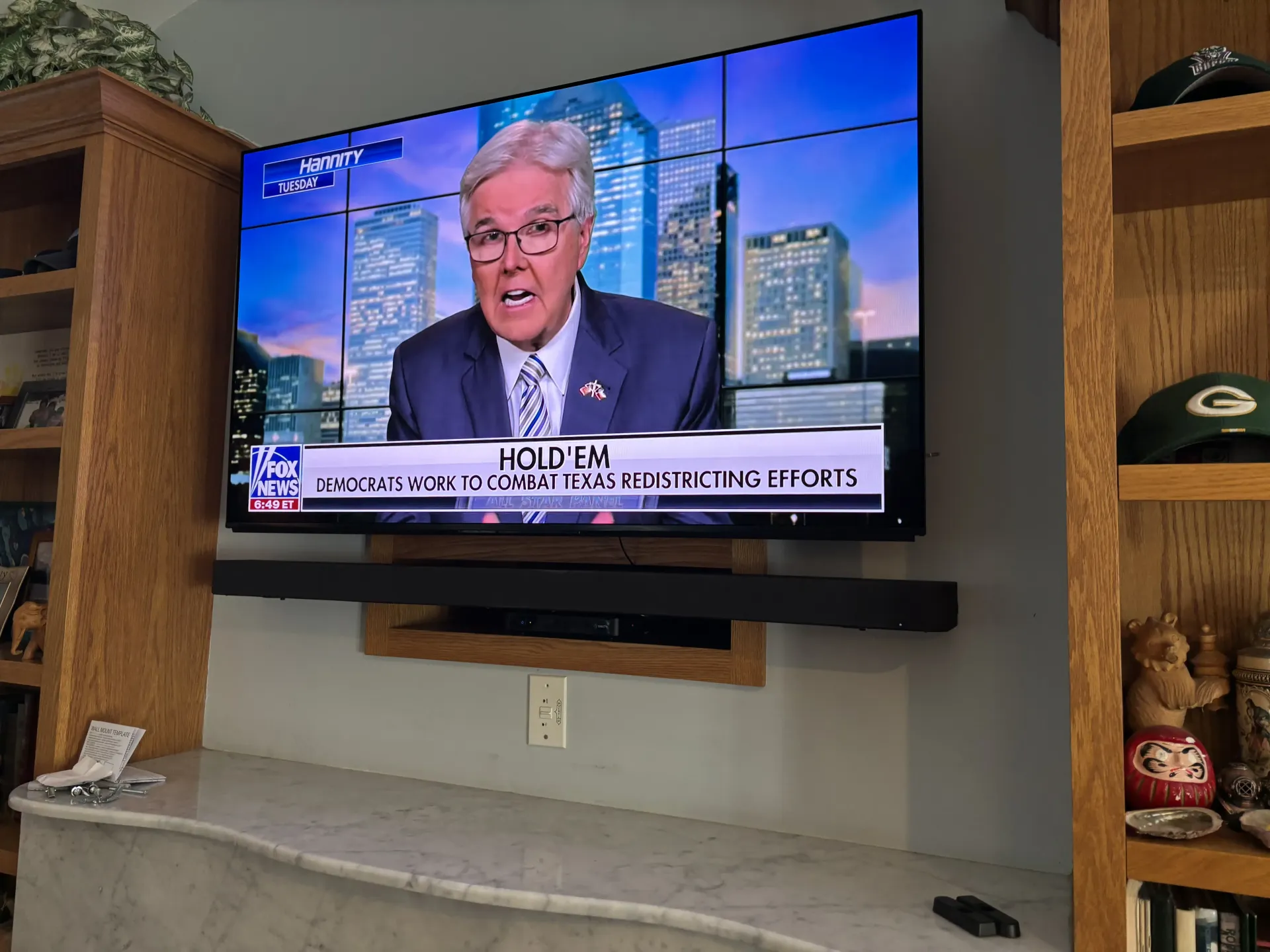 A wall-mounted TV showing a news broadcast above a soundbar and mantel, flanked by wooden bookshelves in a living room.