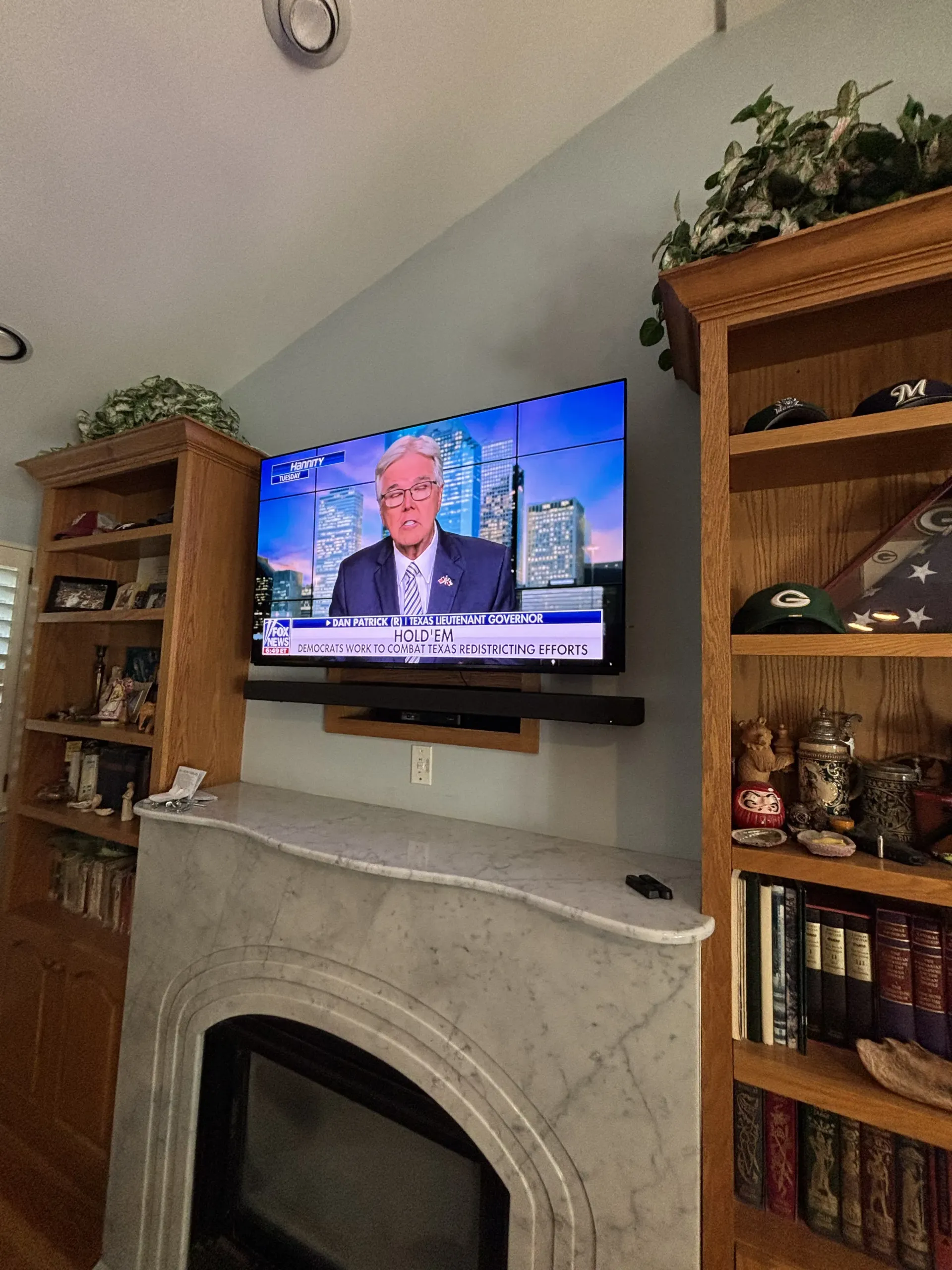 A television mounted above a marble fireplace between two wooden bookshelves filled with collectibles and books.