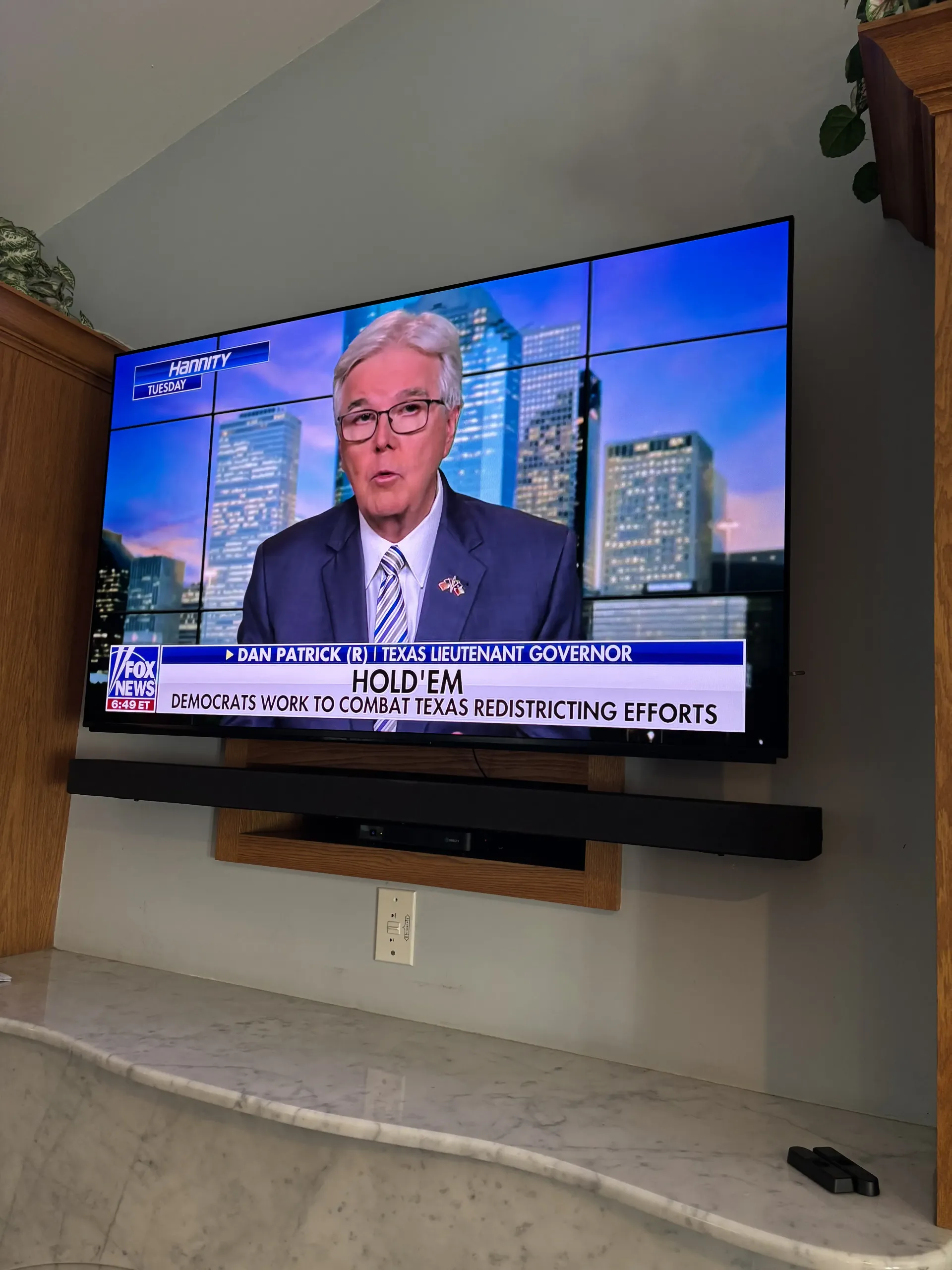A television mounted on a wall above a soundbar and stone mantel, displaying a news program featuring a man in a suit.
