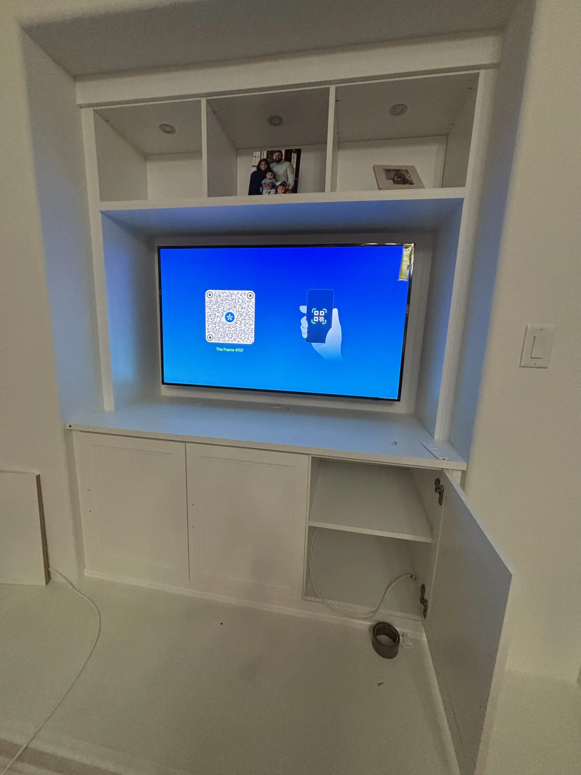 A white built-in wall unit with an open shelf, a TV displaying a setup screen, and storage cabinets with one door open.