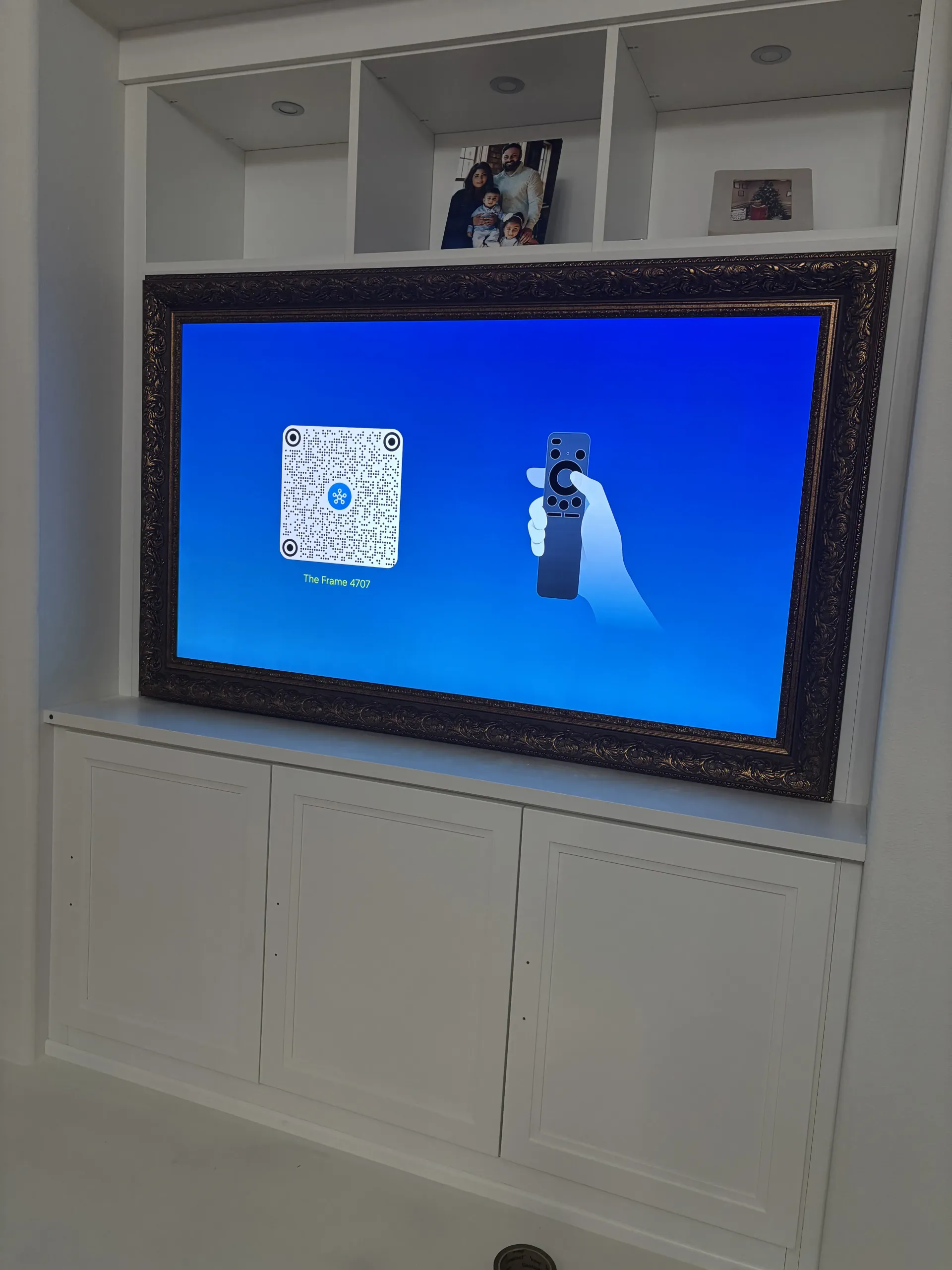 A wall-mounted TV framed in dark wood displays a blue setup screen with a QR code and an illustration of a remote.