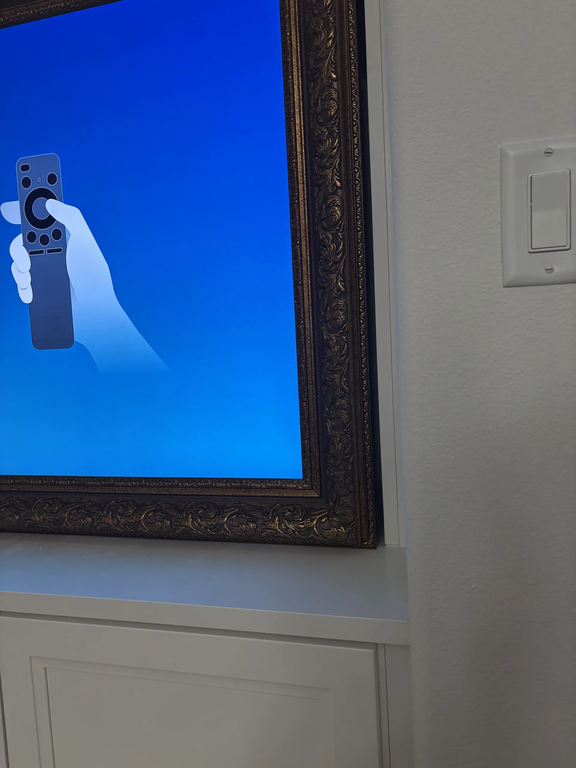 A television screen displaying a remote control icon against a blue background, framed in ornate gold, next to a wall switch.