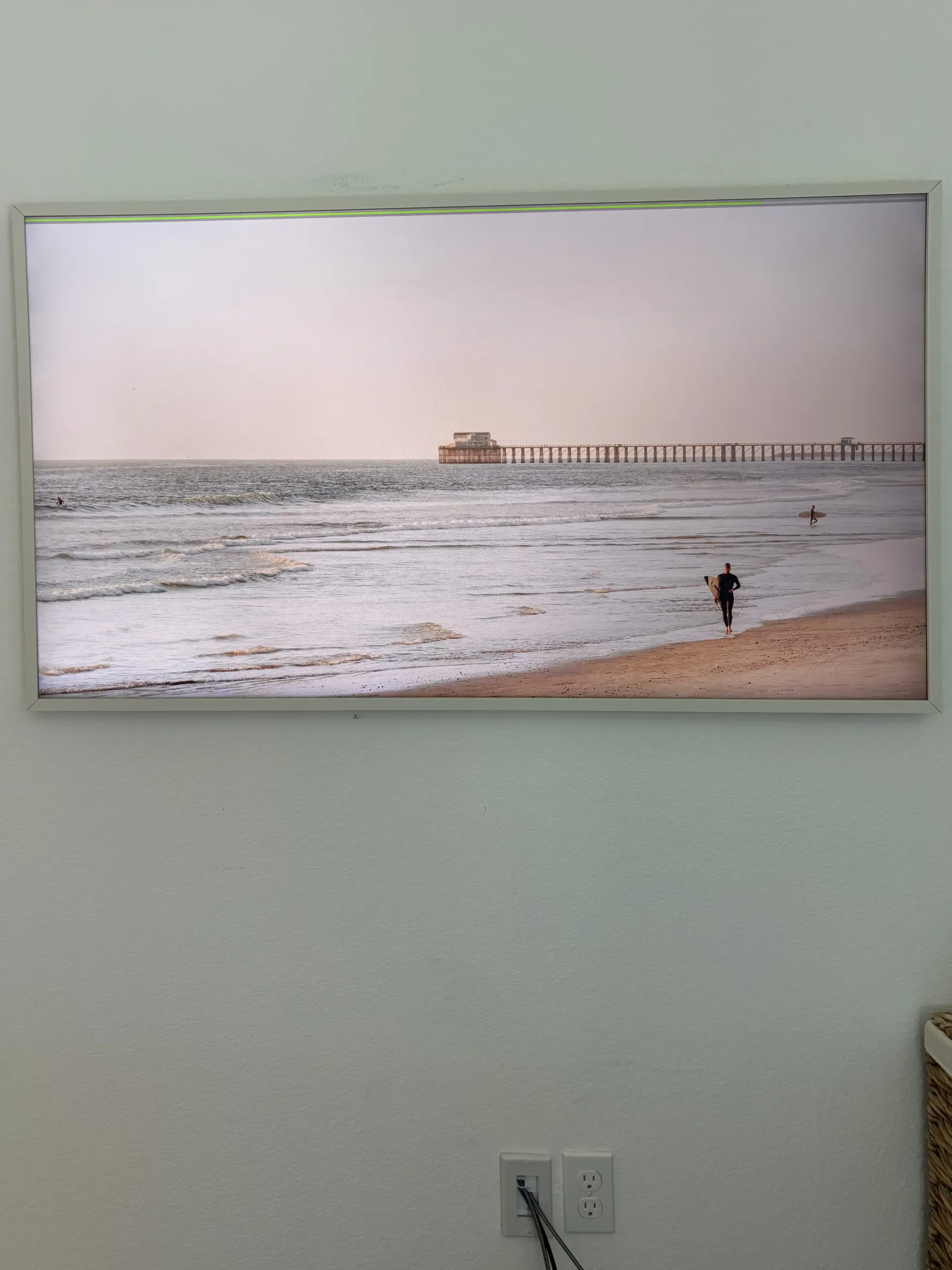 A wall-mounted print of a beach scene with a long pier extending into the ocean and a person standing near the shoreline.