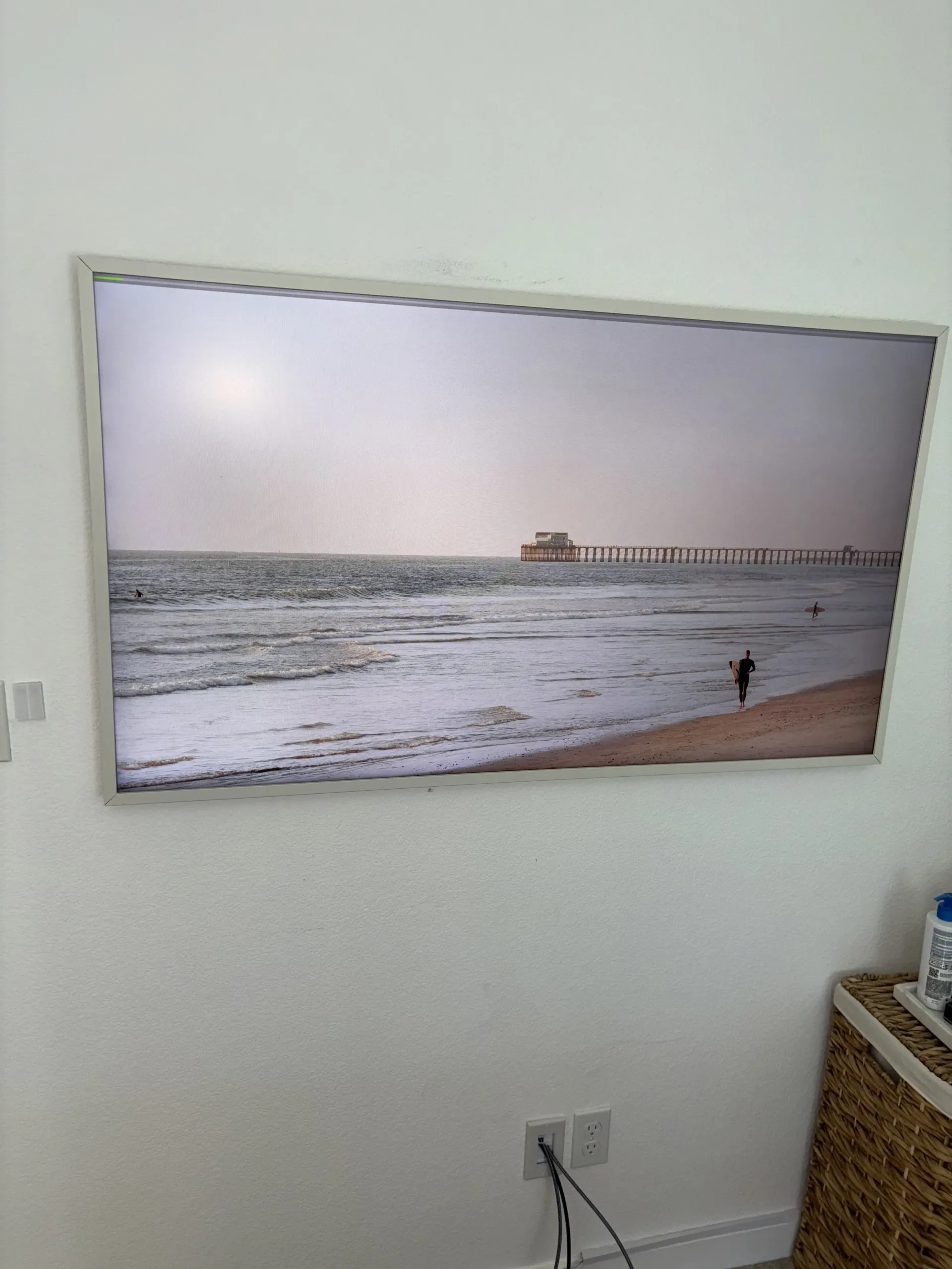 A framed digital display on a white wall showing a beach scene with a pier in the distance and a person on the sand.