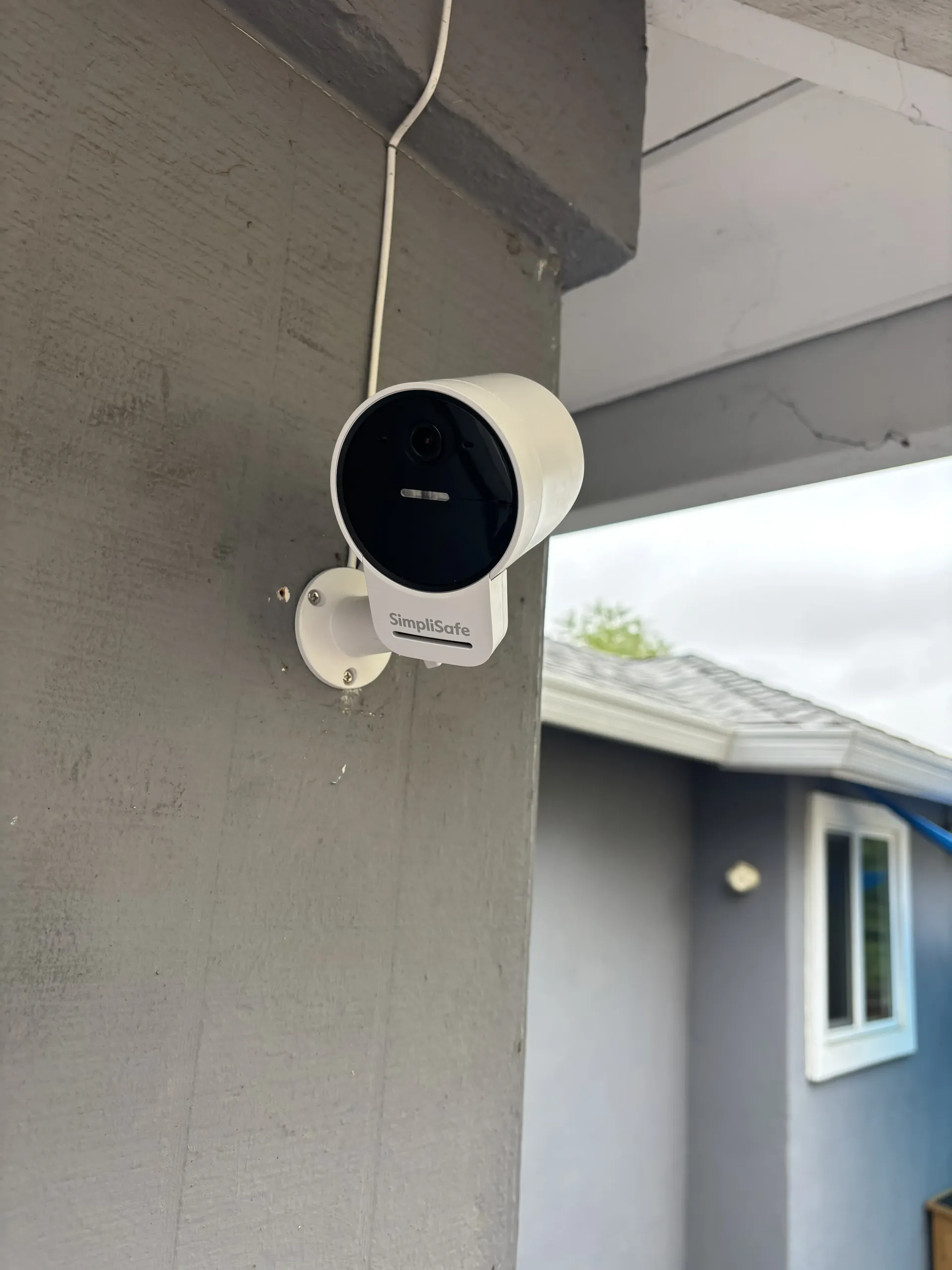 A white Wyze outdoor security camera mounted on a gray exterior wall next to an overhanging roof.