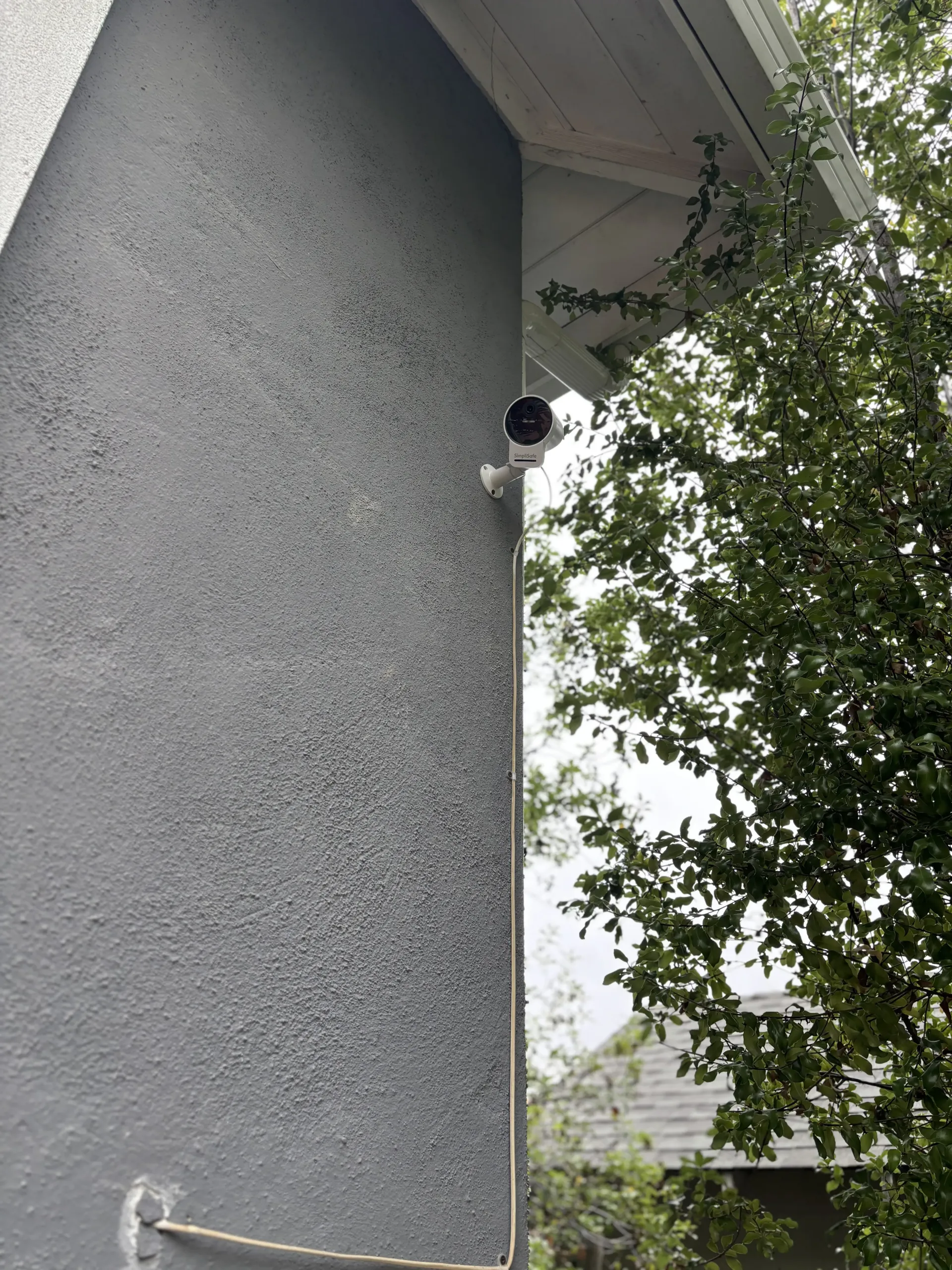 A grey security camera mounted on the side of a grey stucco house with an exposed wire running down the wall.