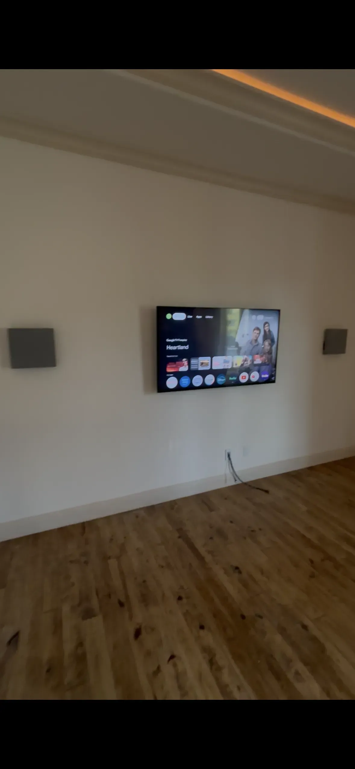 A flat-screen TV mounted on a white wall in a room with hardwood floors, flanked by two square speakers.