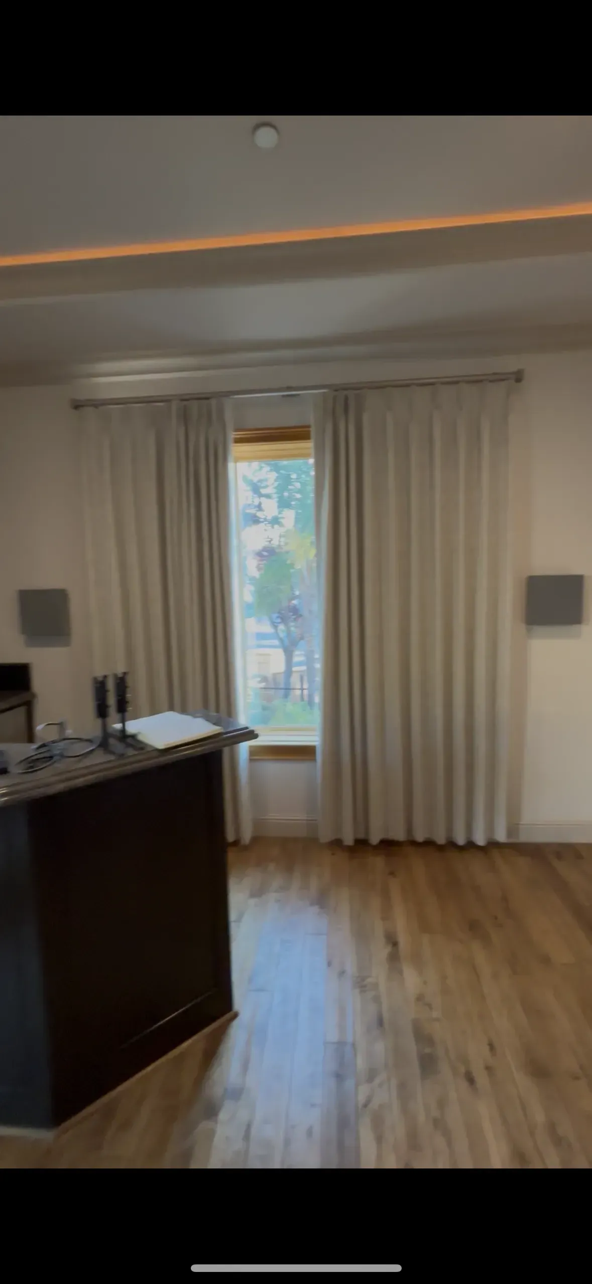 A room with a dark wooden kitchen island in the foreground, light-colored curtains covering a window, and wood flooring.