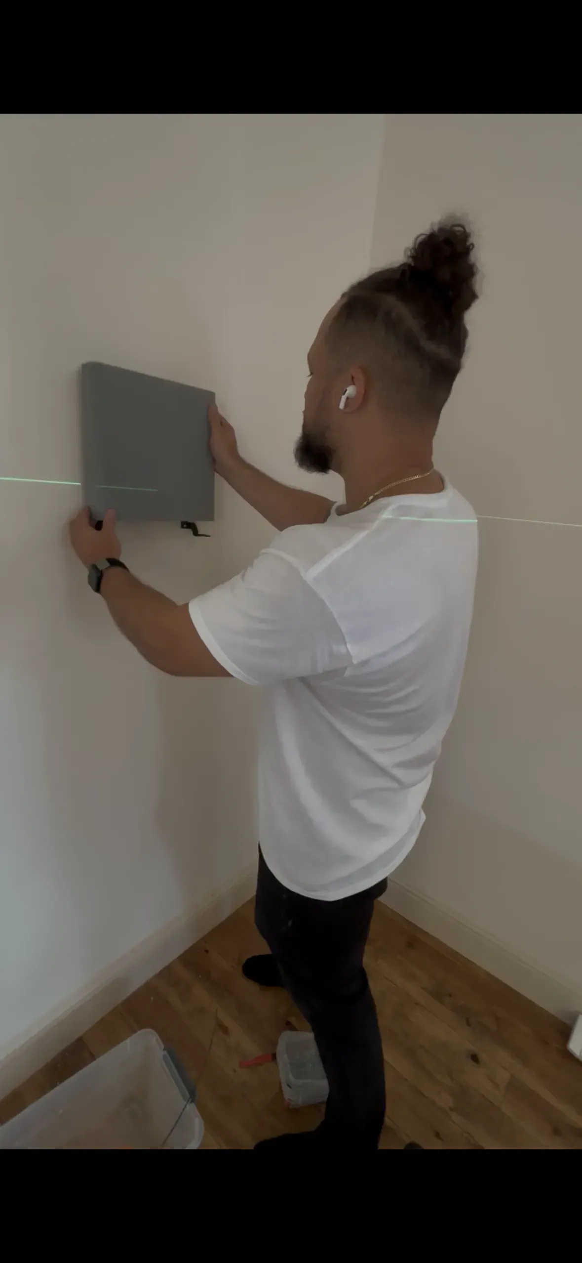 A person wearing a white t-shirt attaches a small, gray rectangular panel to a plain wall in a room with wood flooring.