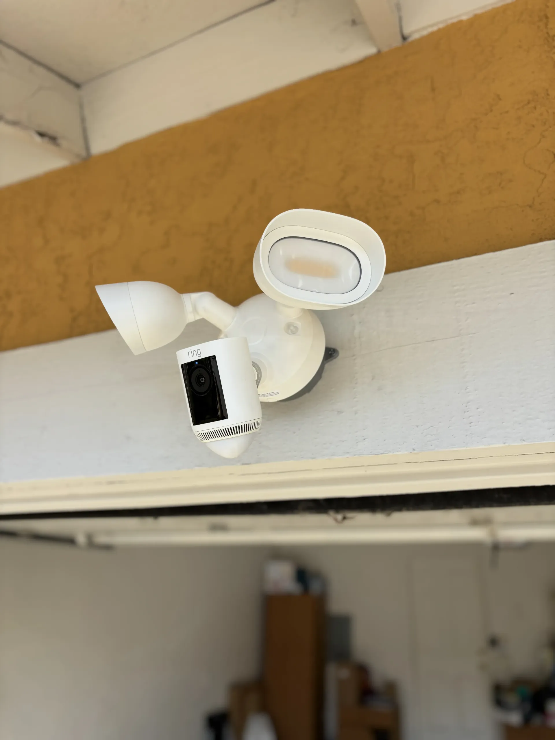A white Ring Floodlight Cam mounted on a light-colored surface under a wooden beam.