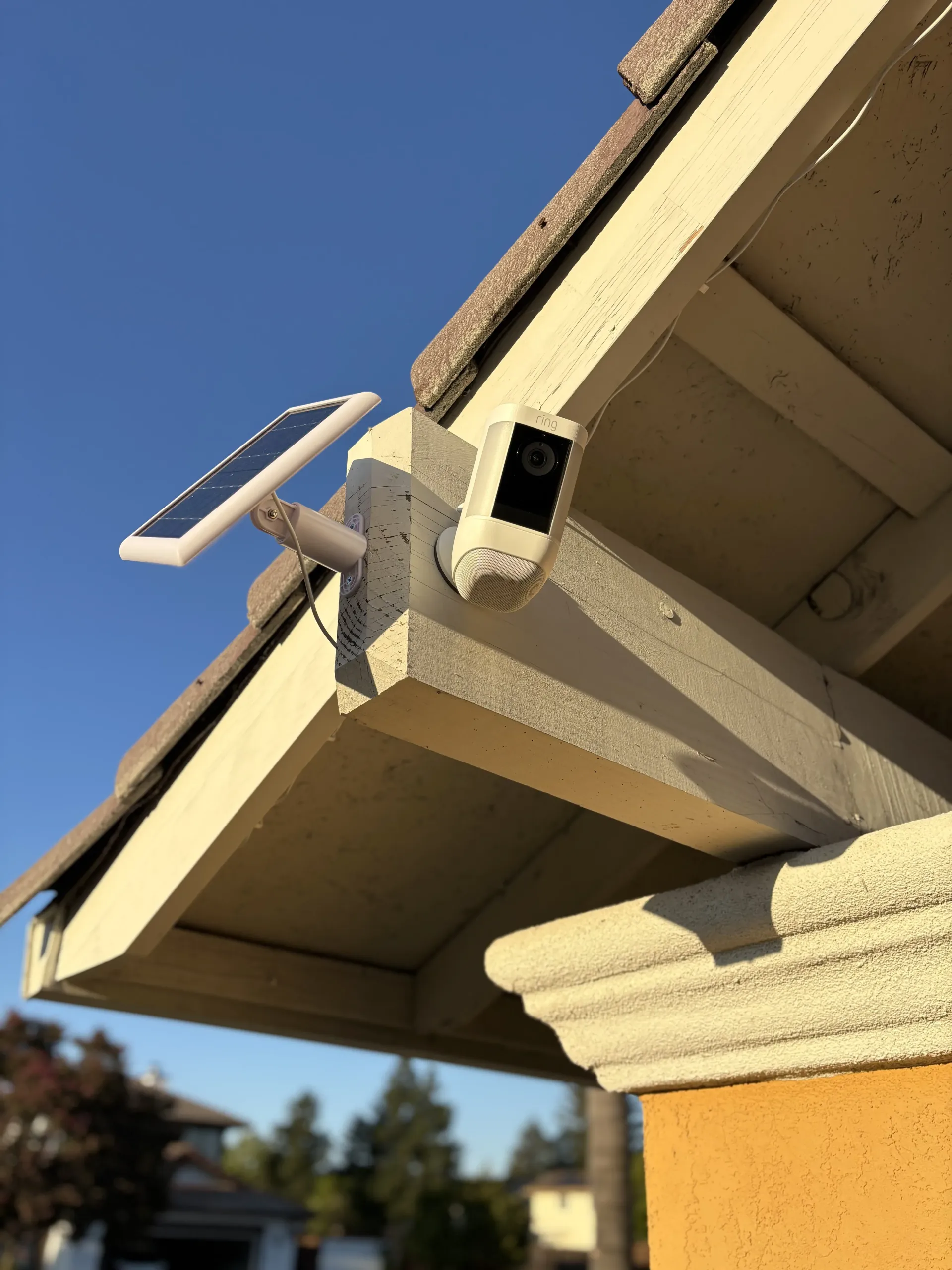 A security camera with an attached solar panel mounted under the eaves of a residential roof against a clear blue sky.